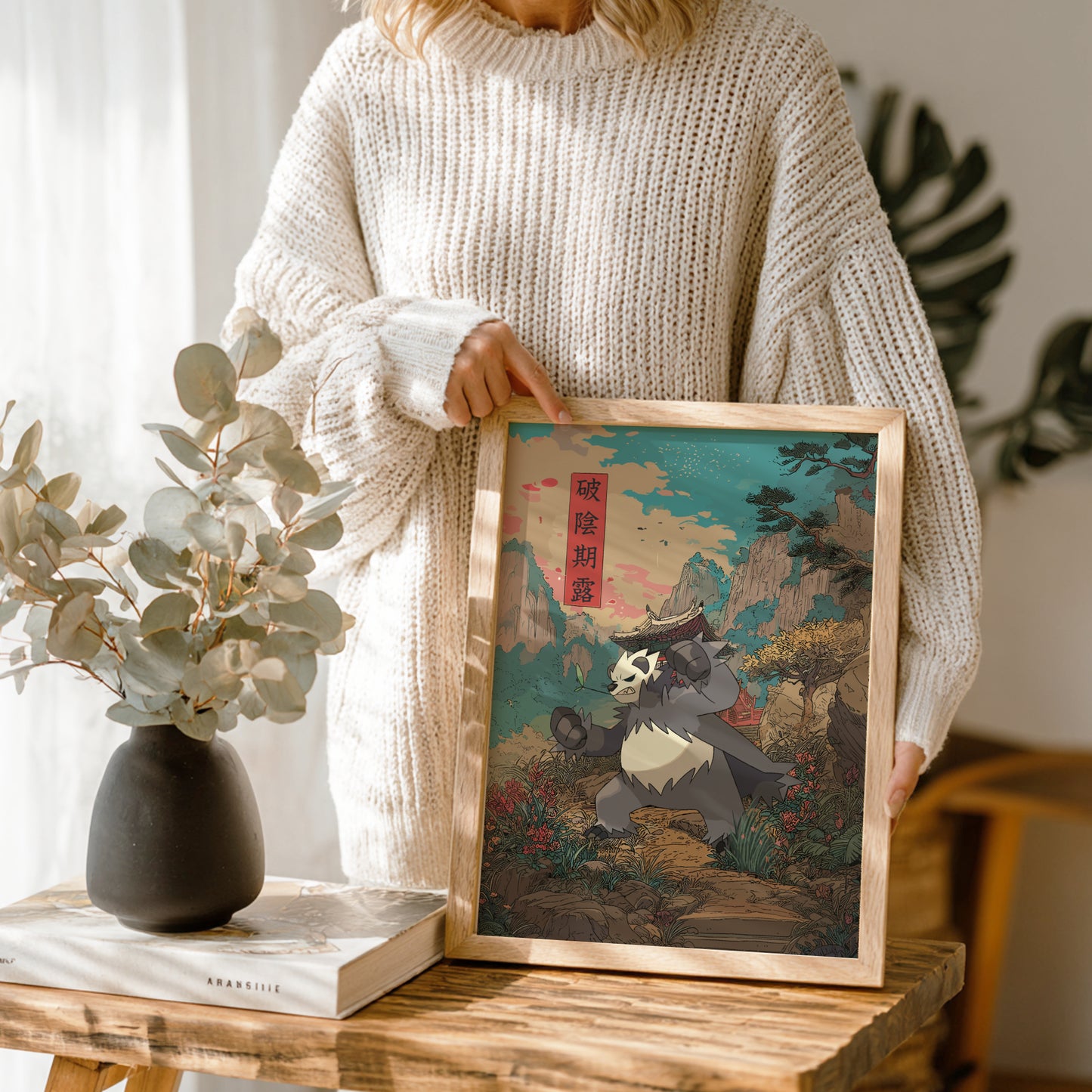 Person holding a framed artwork of Pangoro Pokemon in a scenic setting, with a vase and book on a wooden table.