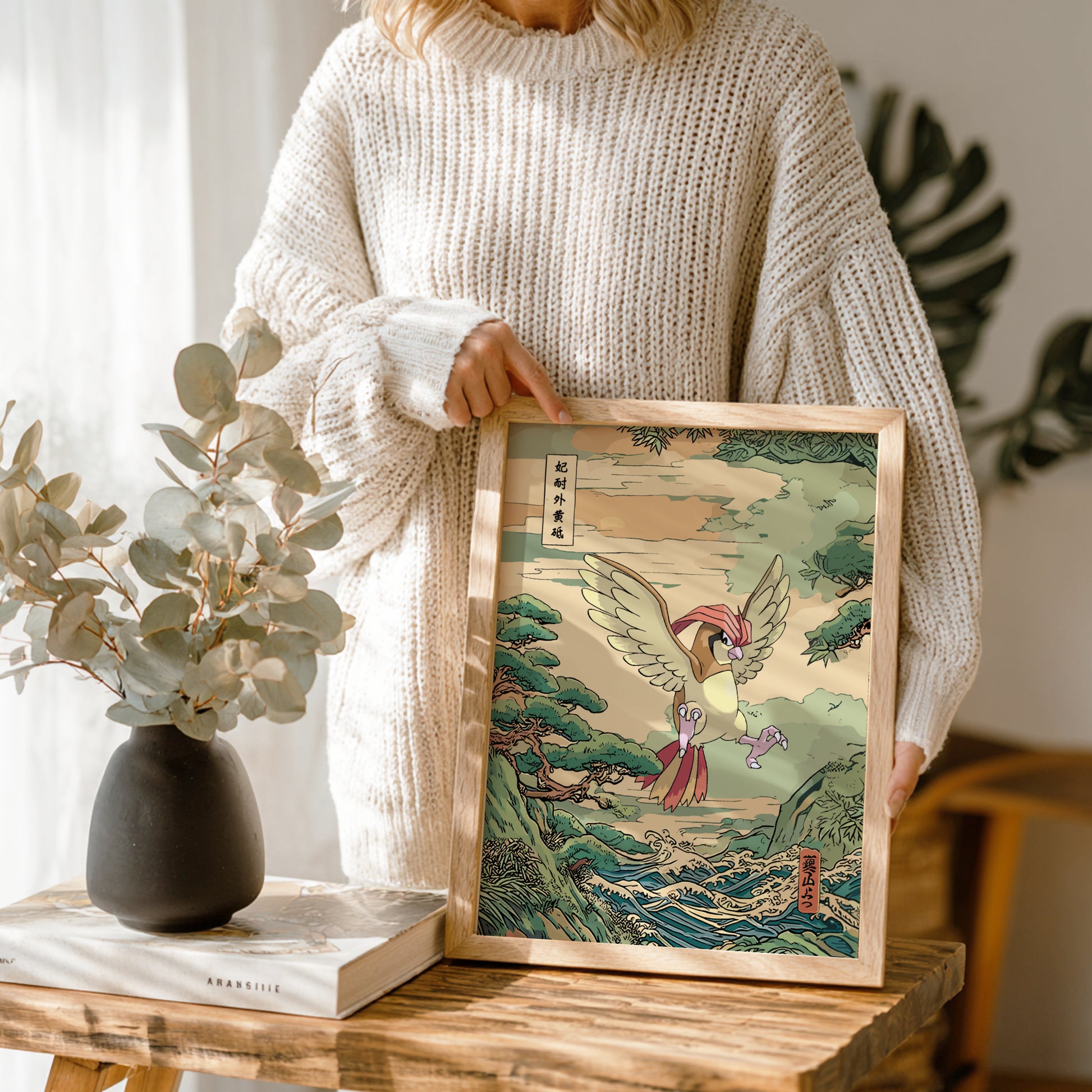 Person holding a framed artwork of Pidgeotto Pokemon with a nature-themed design, standing next to a vase and books on a wooden table.