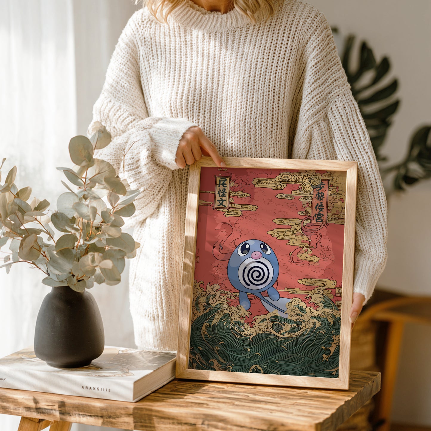 Person holding a framed artwork with Poliwag Pokemon character on a red background, standing next to a vase and books.