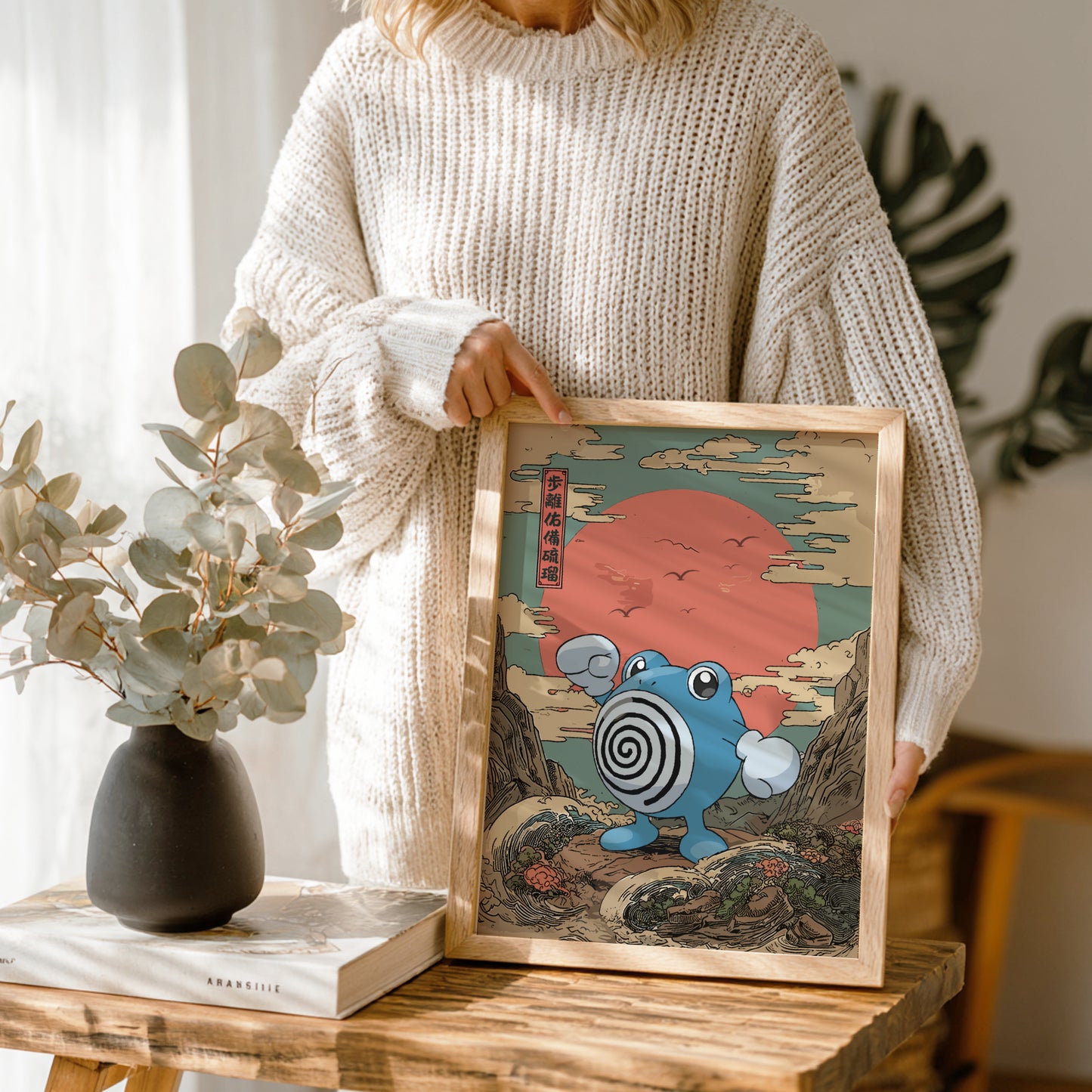 Person holding a framed artwork with Poliwhirl Pokemon and Japanese text, standing in a room with plants and decor.