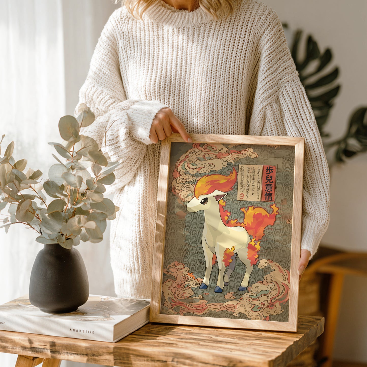 Person holding a framed artwork of Ponyta Pokemon on a wooden table with a vase and book in the background.