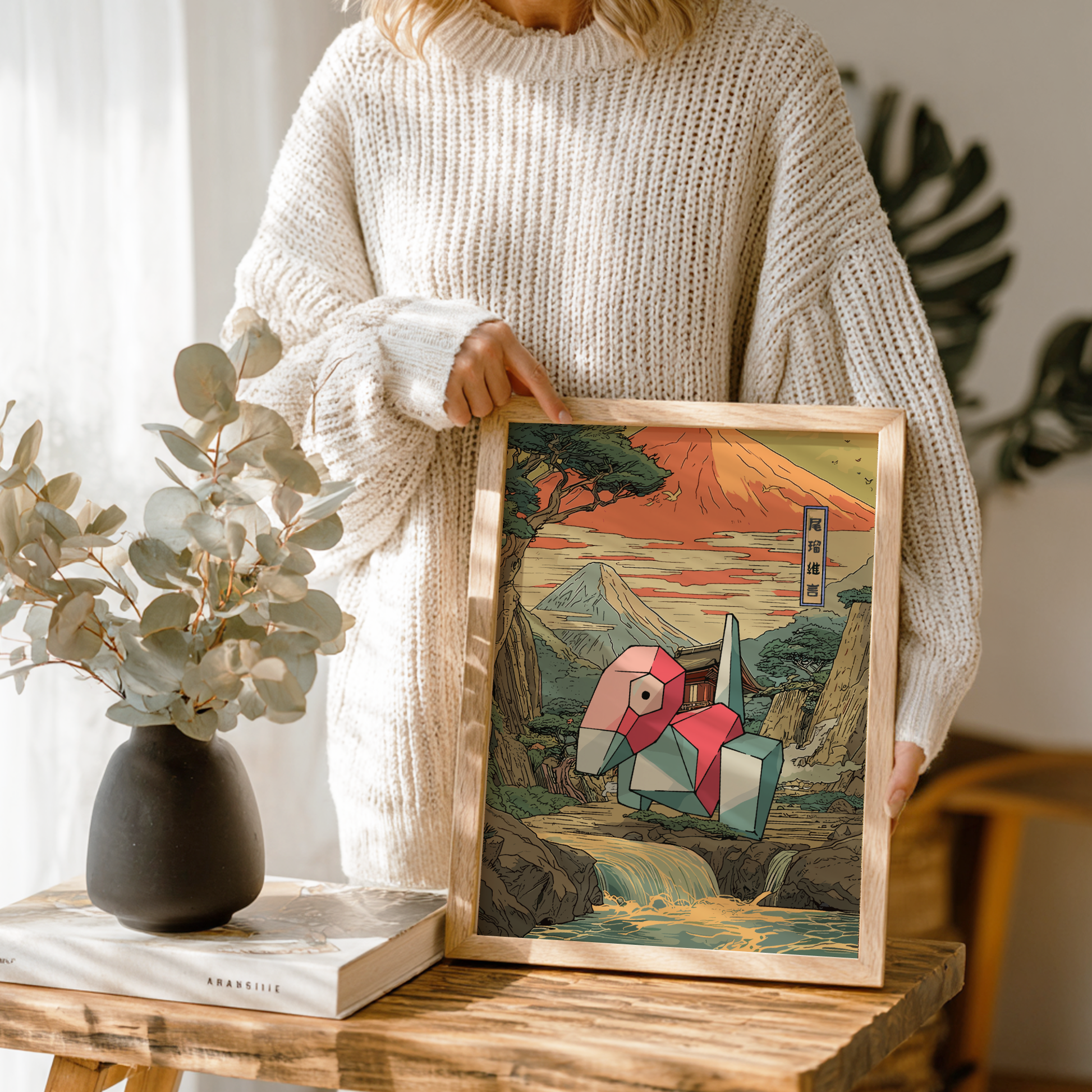 Person holding a framed artwork with a scenic design and Porygon Pokemon, standing in a room with a vase and book on a table.
