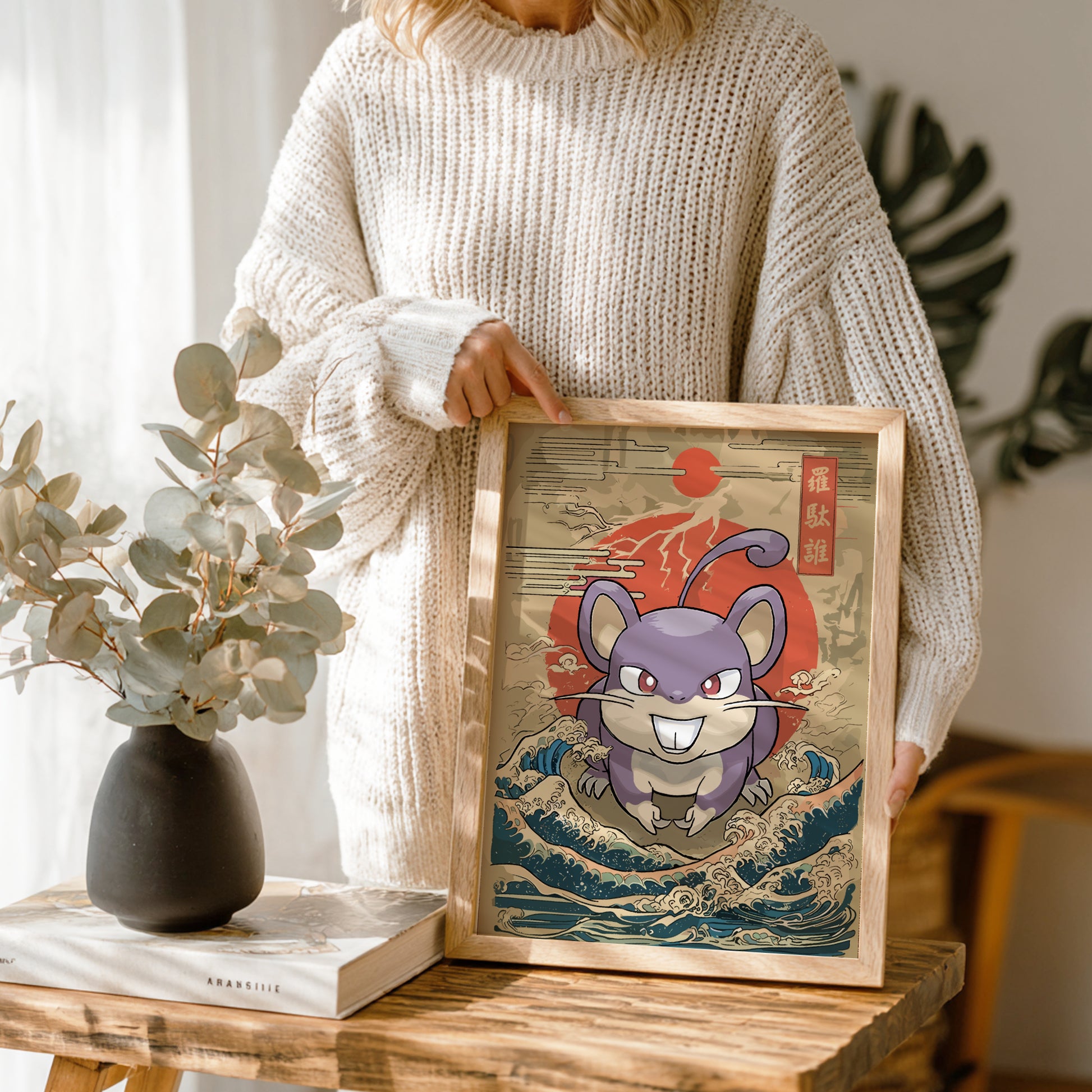 Person holding a framed artwork of a stylized Rattata Pokemon on a wooden table with a vase and book in the background.