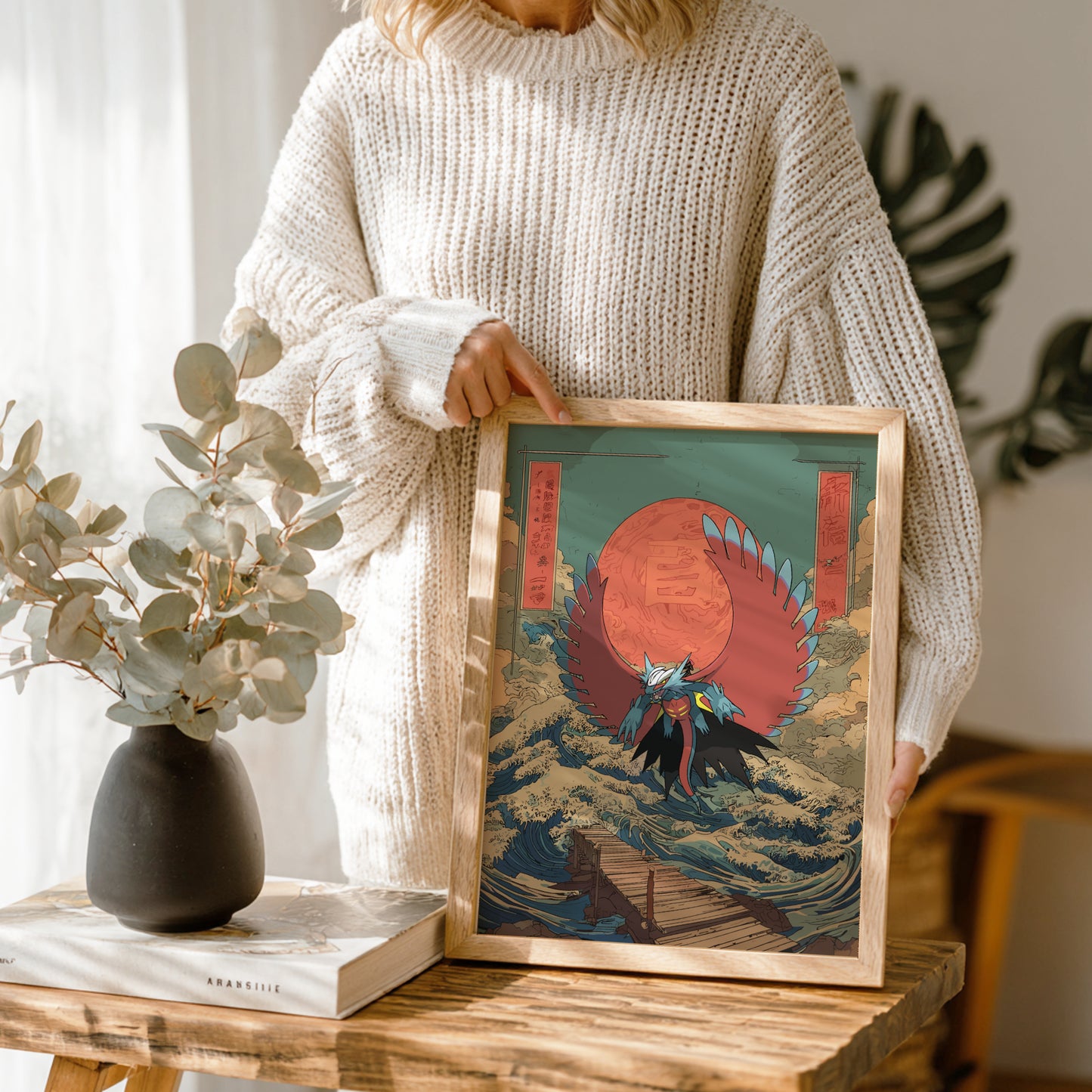 Person holding a framed artwork of Roaring Moon Pokemon against a red sun, with a vase and book on a wooden table.