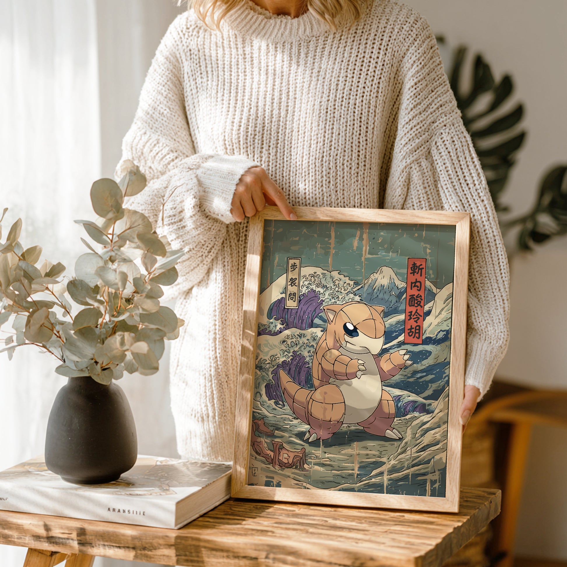 Person holding a framed artwork of Sandshrew Pokemon in a scenic setting, with a vase and books on a table.