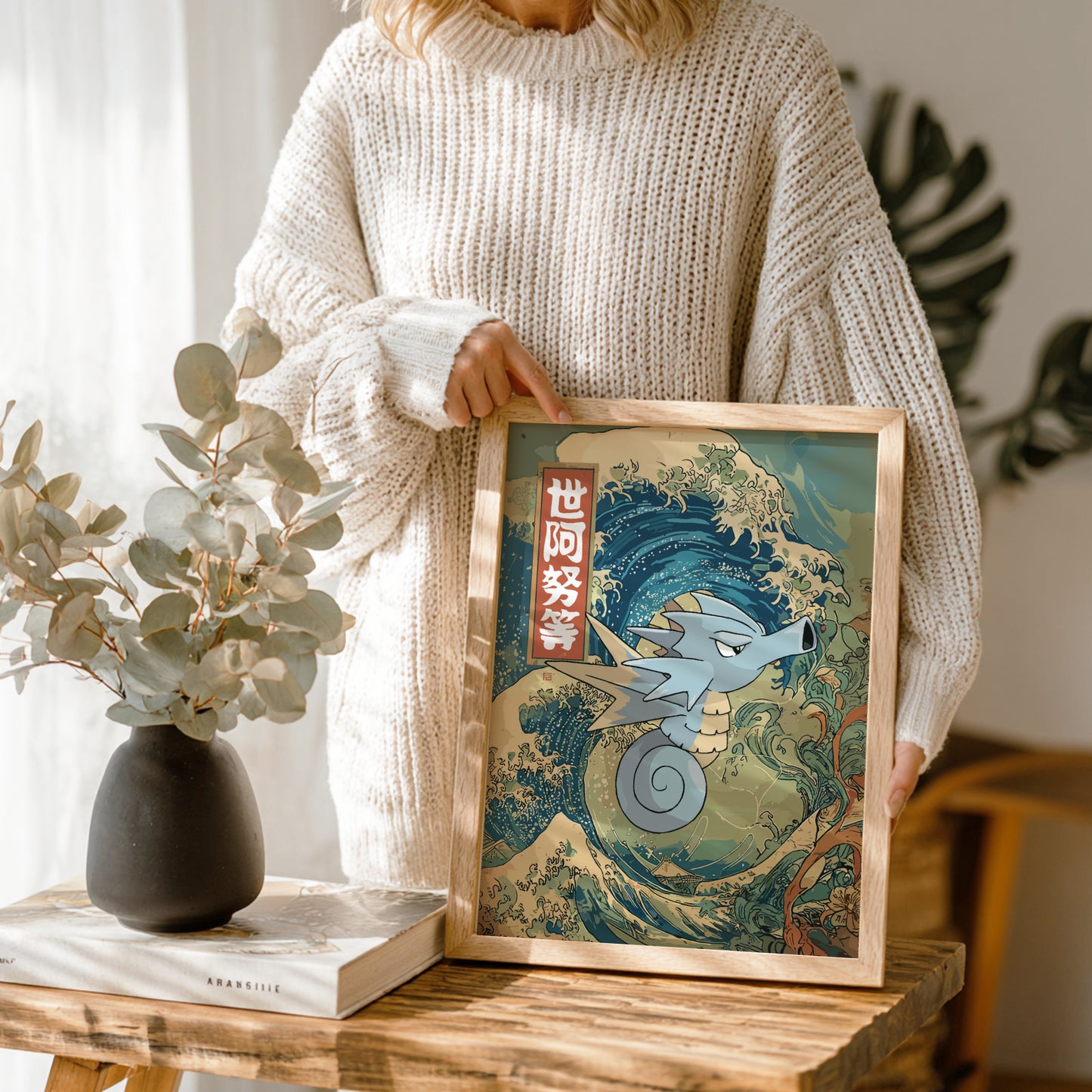Person holding a framed artwork of a wave with Seadra Pokemon, standing in a room with a vase and book on a table.