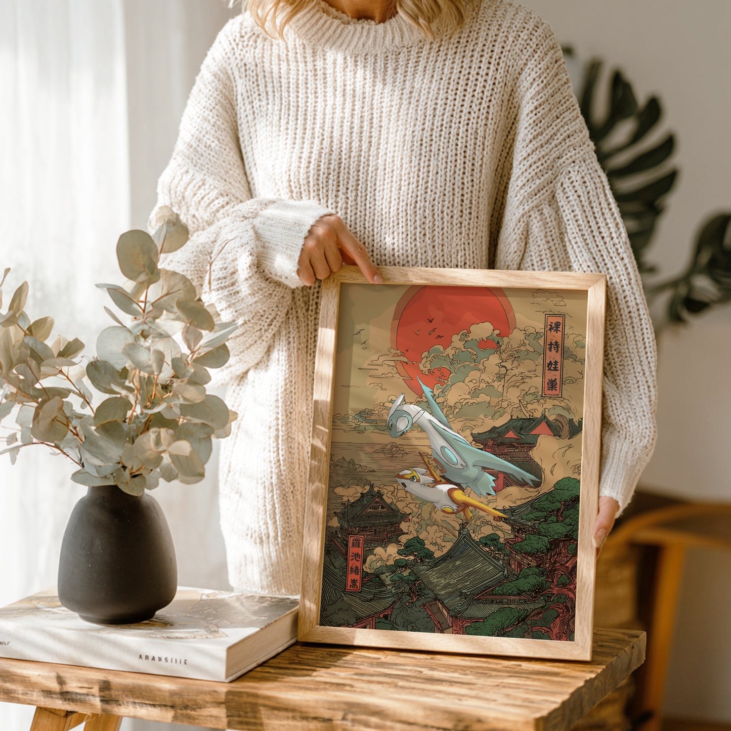 Person holding a framed artwork of Shiny Latias and Latios Pokemon with a scenic design, standing in a room with a vase and books on a table.
