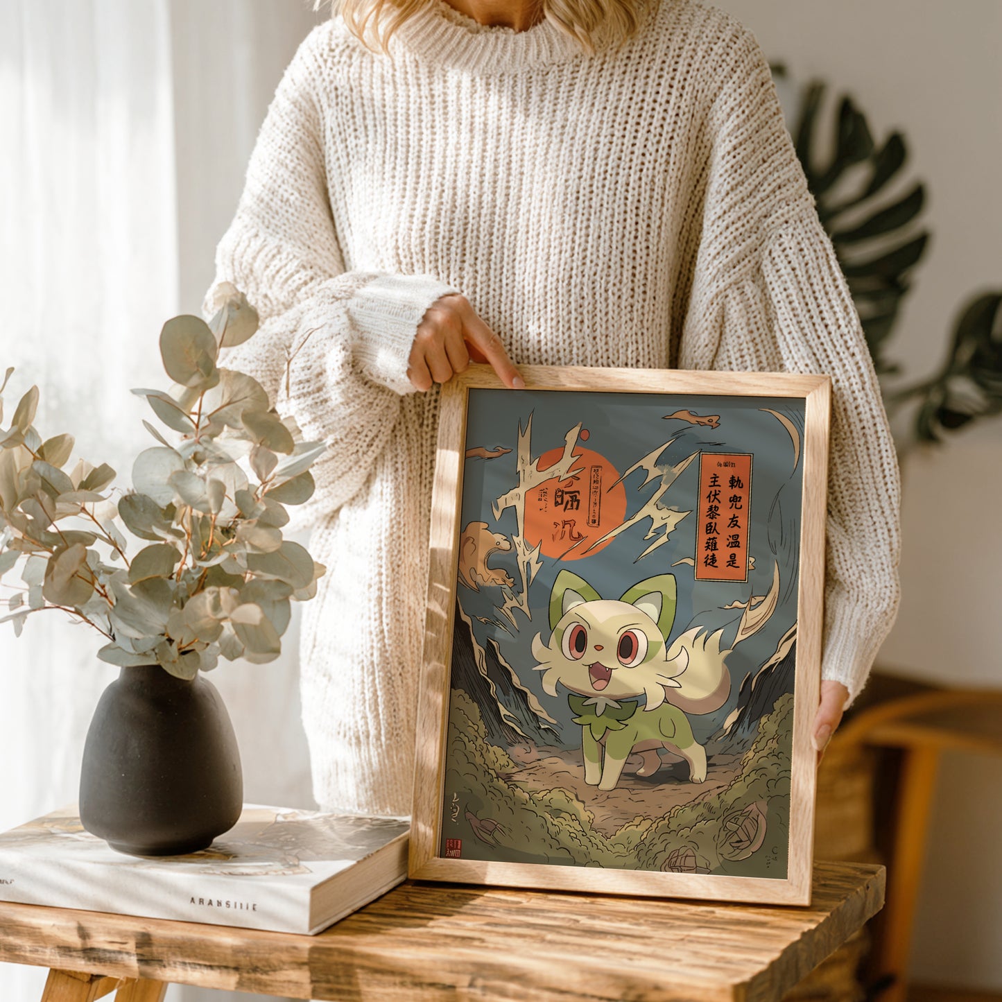 Person holding a framed illustration of Sprigatito Pokemon character on a wooden table with a vase and book in the background.