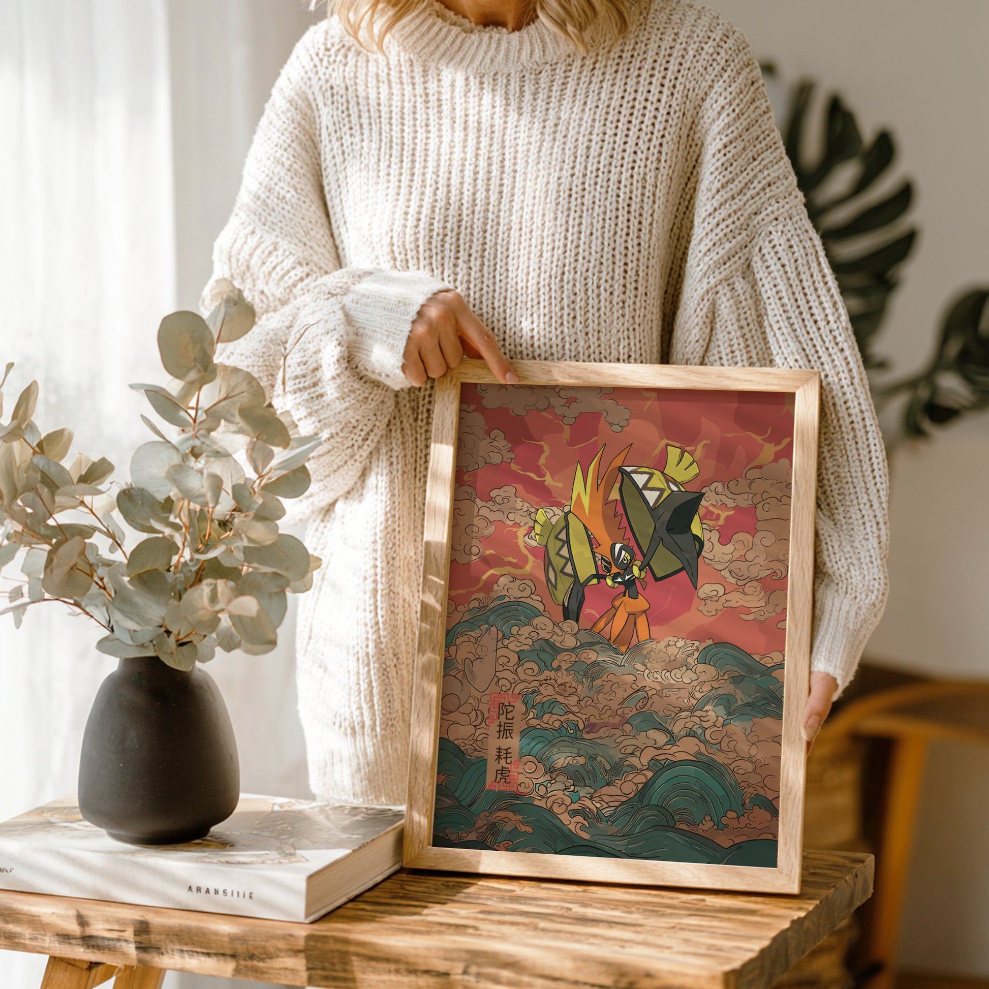Person holding a framed artwork with a colorful design of Tapu Koko Pokemon, standing in a room with a vase and book on a table.