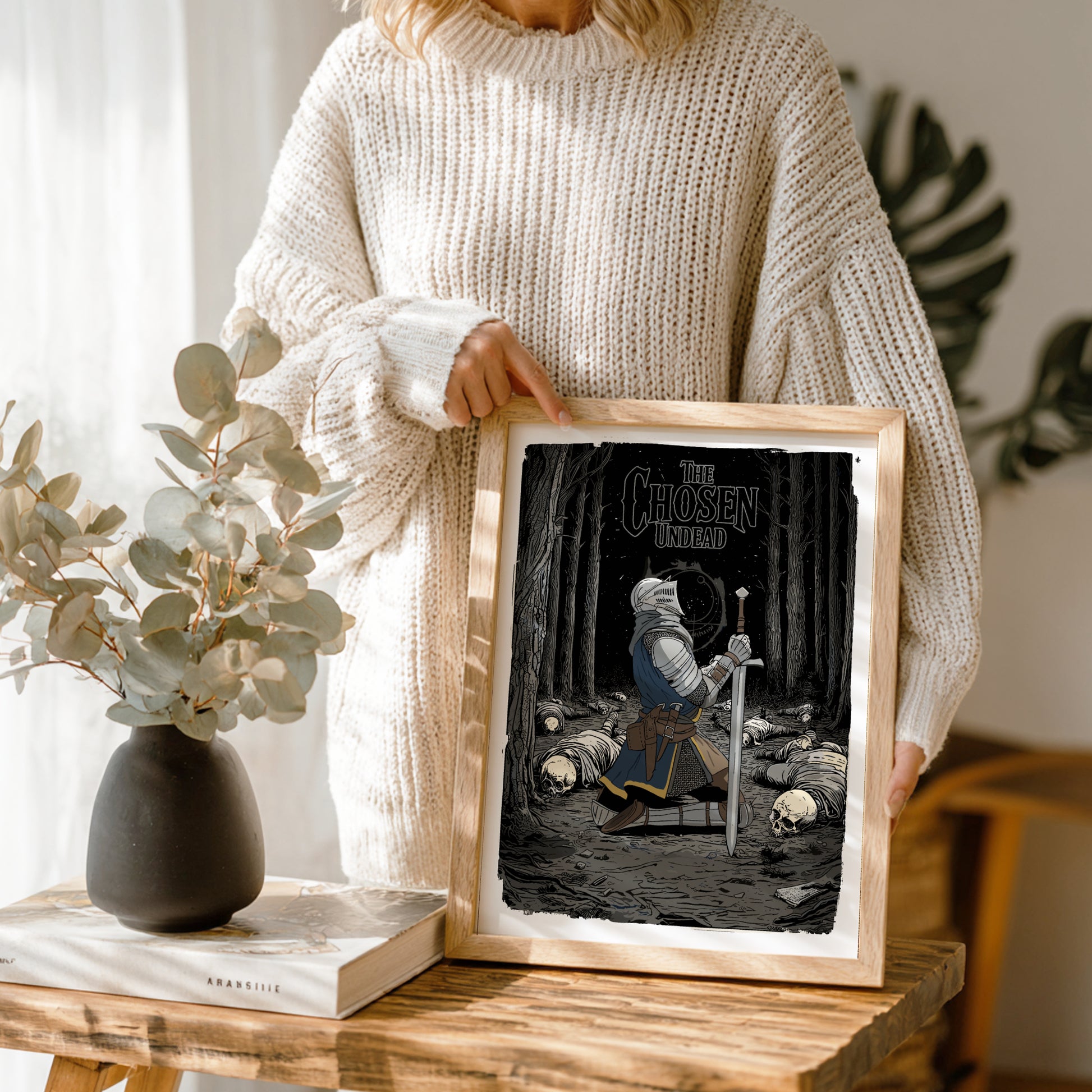 Person holding a framed artwork of The Chose Undead from Dark Souls on a wooden table with a vase and books in the background.