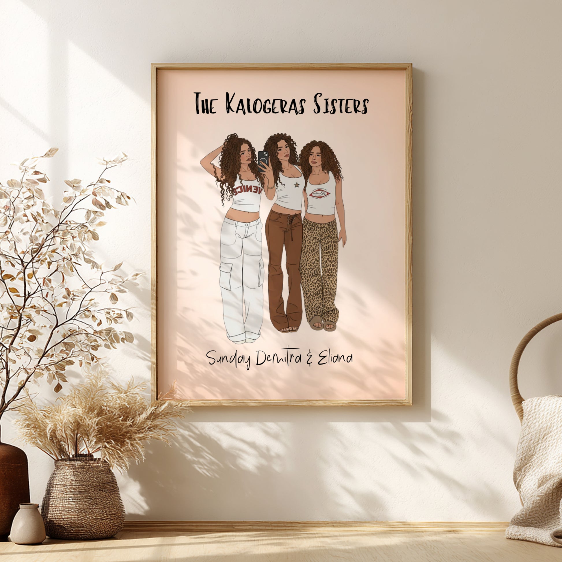 Framed artwork of 'The Kalogeras Sisters' with three illustrated women on a pink background, displayed in a room with decorative elements.