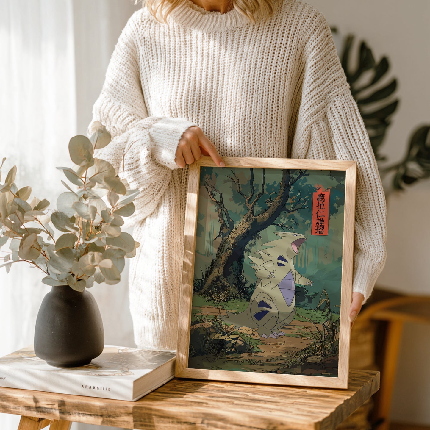 Person holding a framed artwork of a Tyranitar Pokemon in a natural setting, with a vase and book on a table.