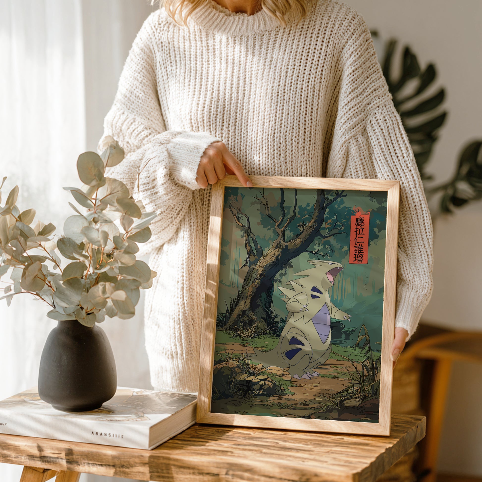 Person holding a framed artwork of a Tyranitar Pokemon in a natural setting, with a vase and book on a table.