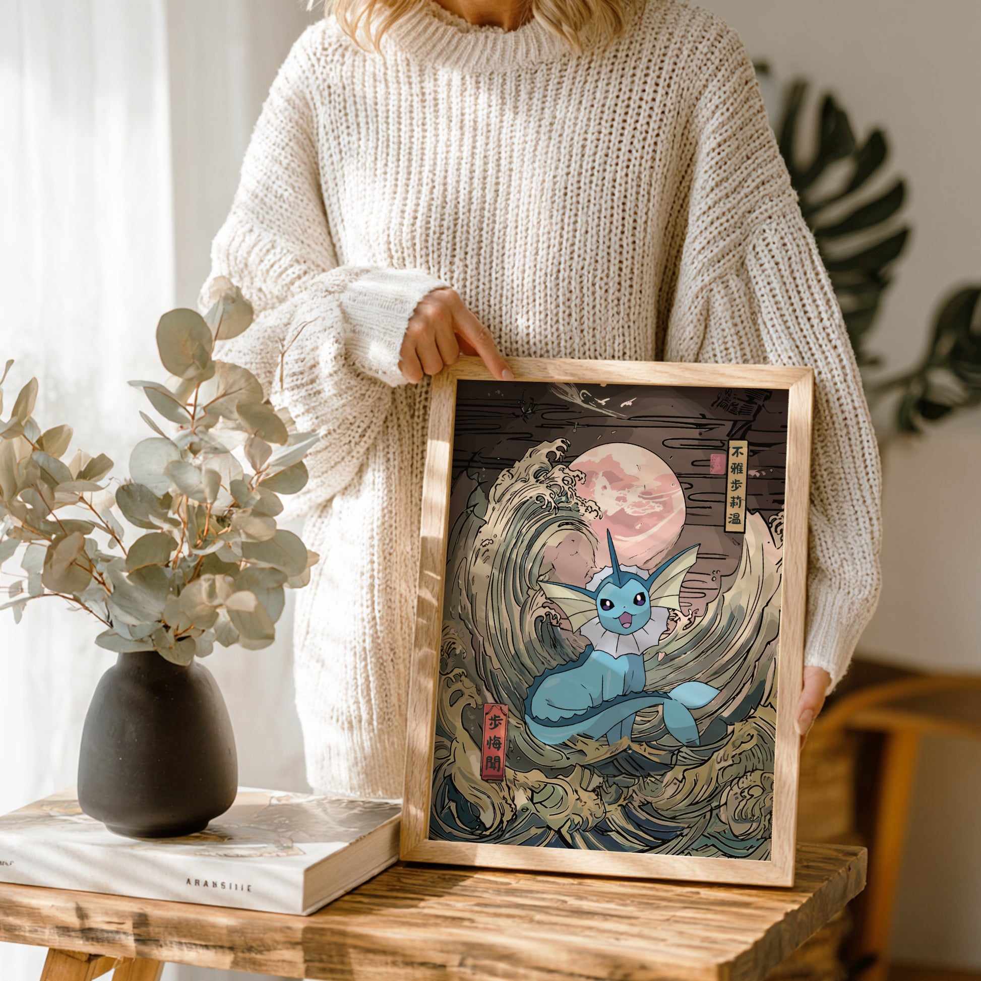 Person holding a framed artwork of Vaporeon Pokemon in a wave, with a vase and book on a wooden table.