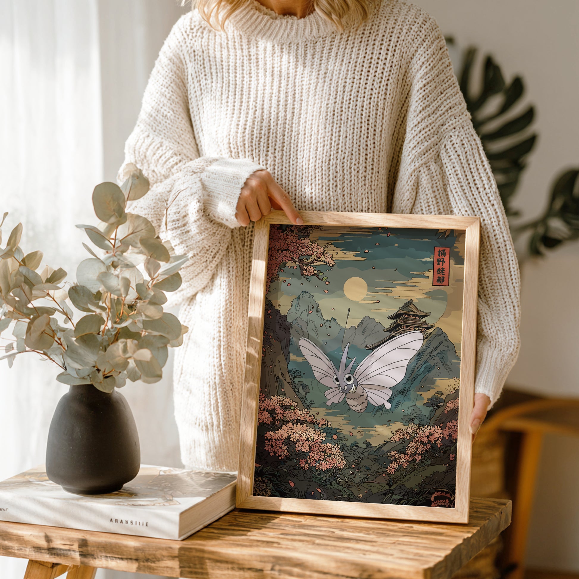 Person holding a framed artwork with Venomoth Pokemon and mountain landscape, next to a vase and books on a wooden table.