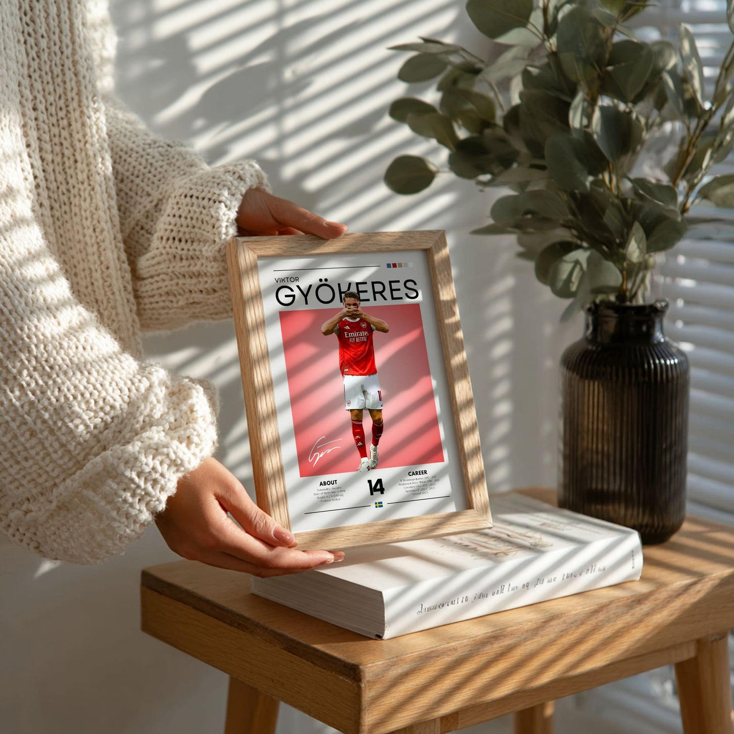 Person holding a framed picture of a person in a red jersey on a wooden table with a plant in the background.