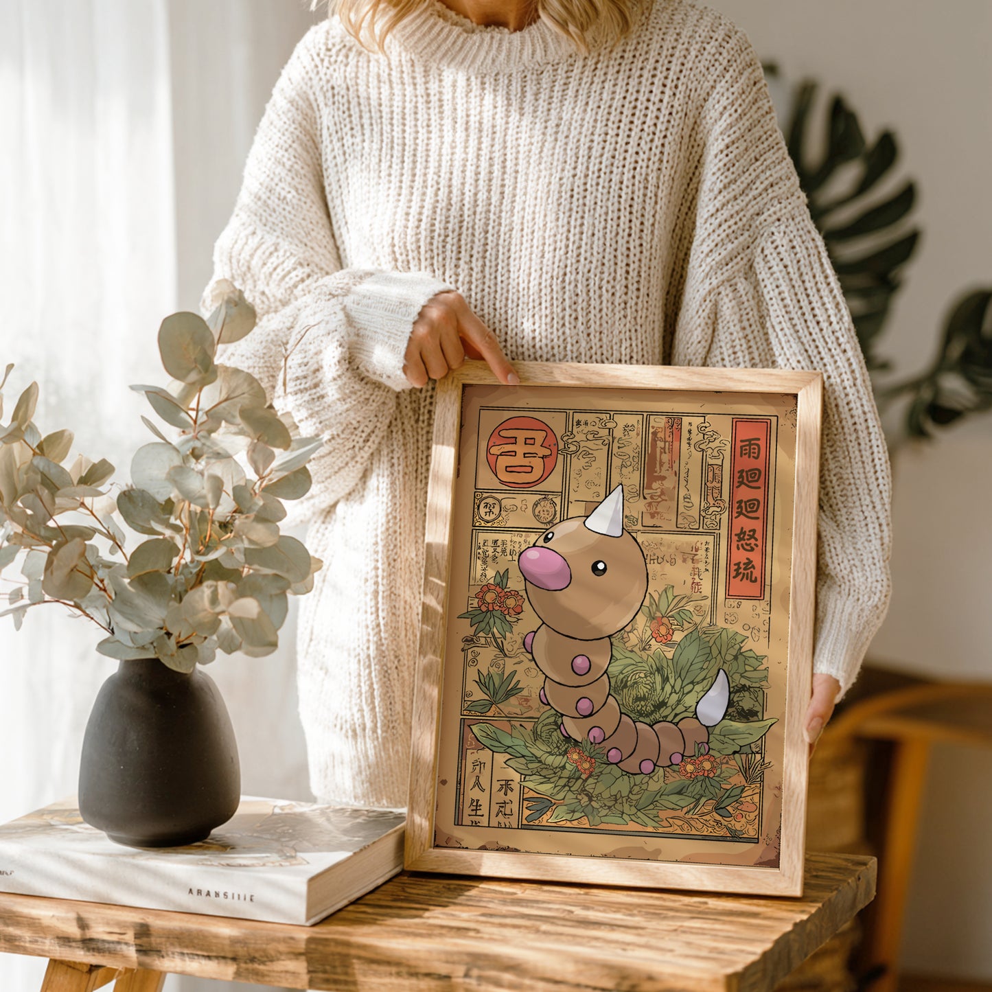 Person holding a framed illustration of Weedle Pokemon on a wooden table.