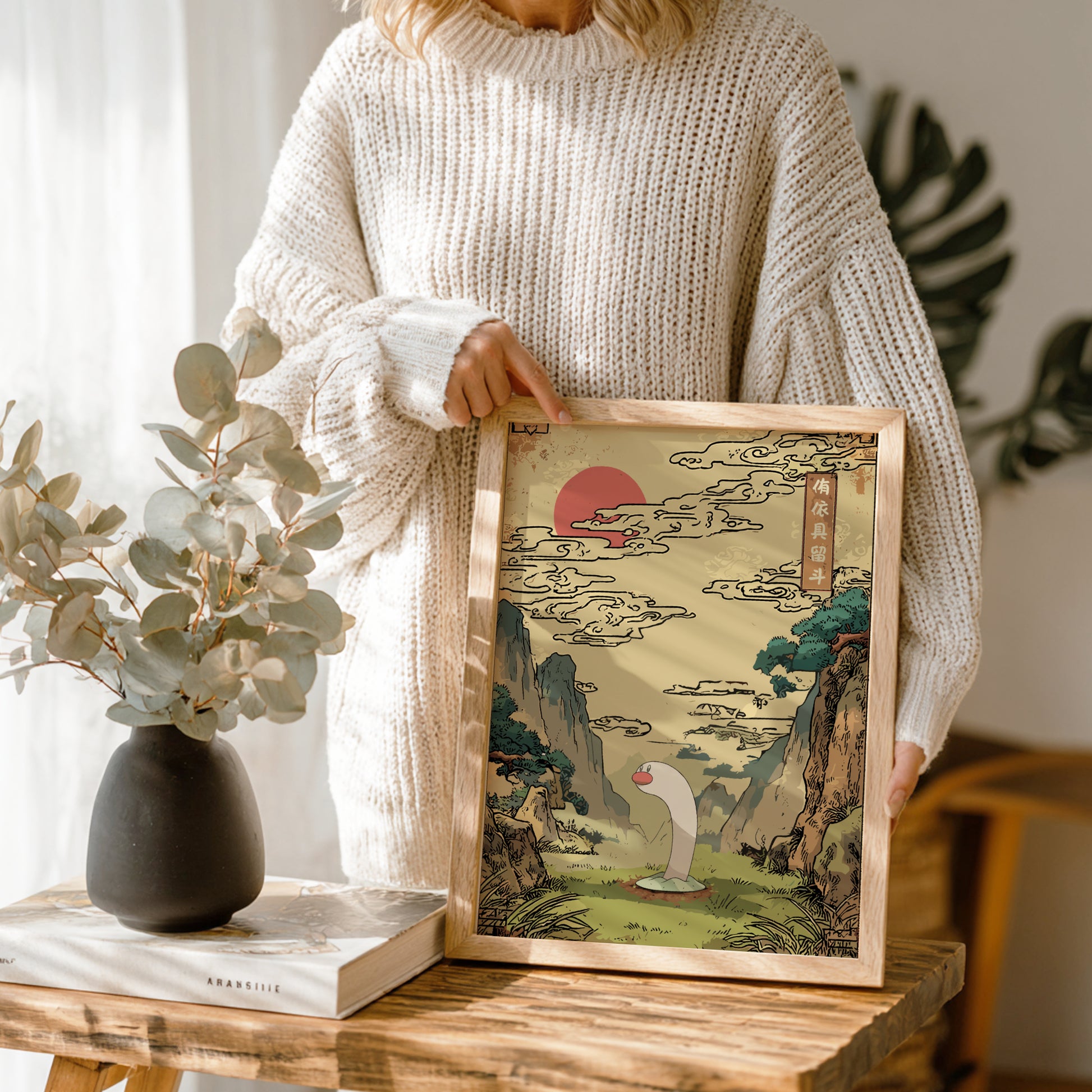 Person holding a framed artwork of Wiglett Pokemon and a landscape with a red sun, sitting at a wooden table with a vase and book.