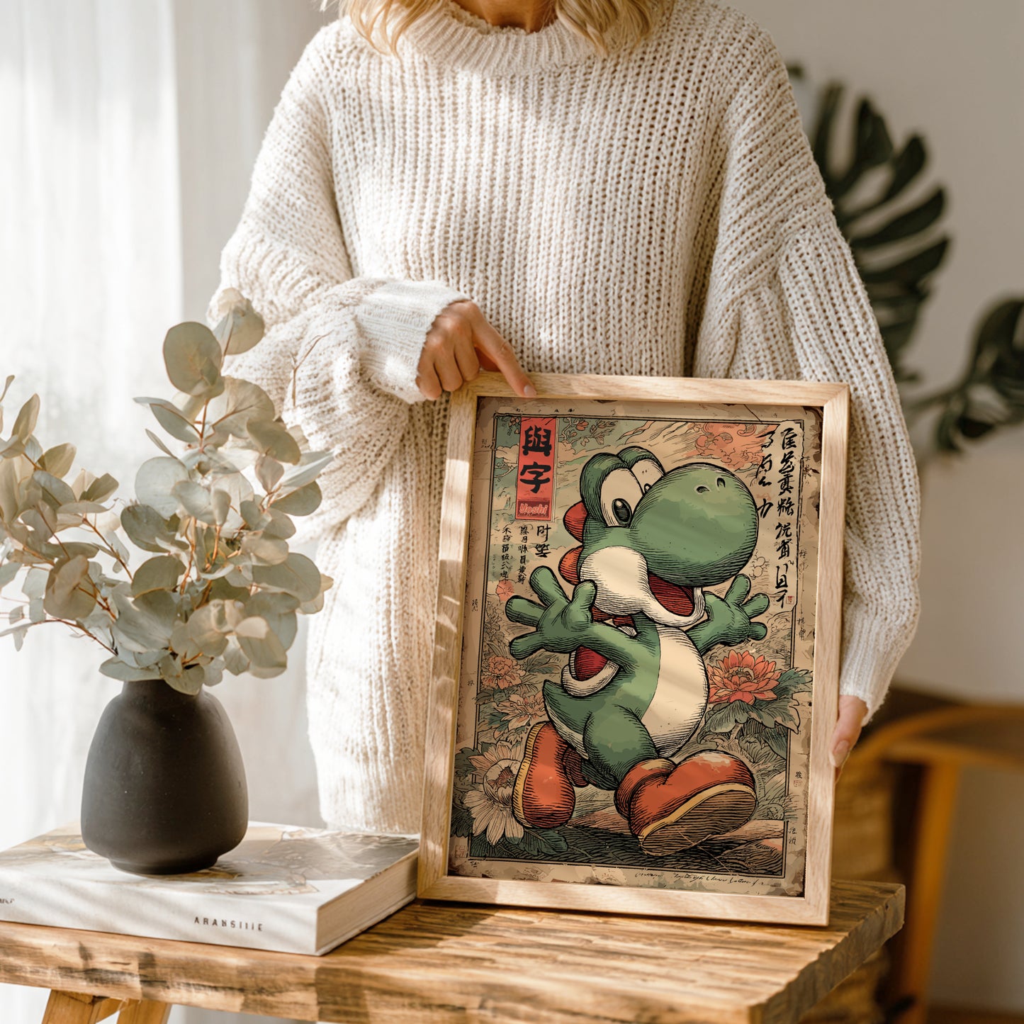 Person holding a framed picture of Yoshi character on a wooden table with a vase and books in the background.