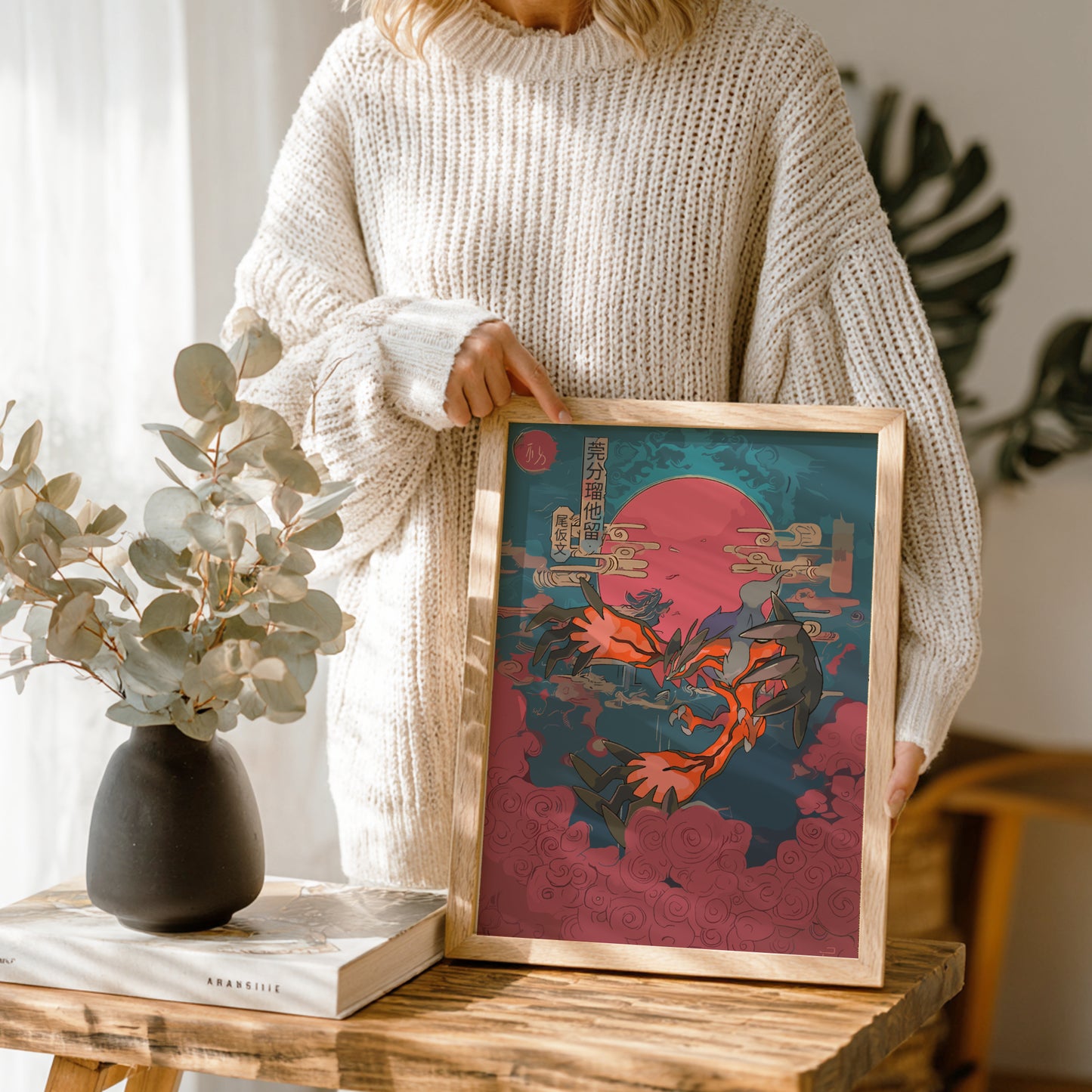 Person holding a framed artwork of Yveltal Pokemon on a wooden table with a vase and book in the background.