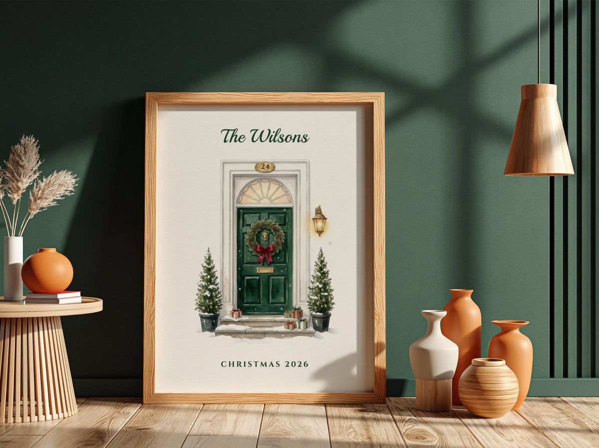 A framed personalized Christmas art print leaning against a dark green wall on a wooden floor, surrounded by decorative vases, a side table, and a hanging lamp.