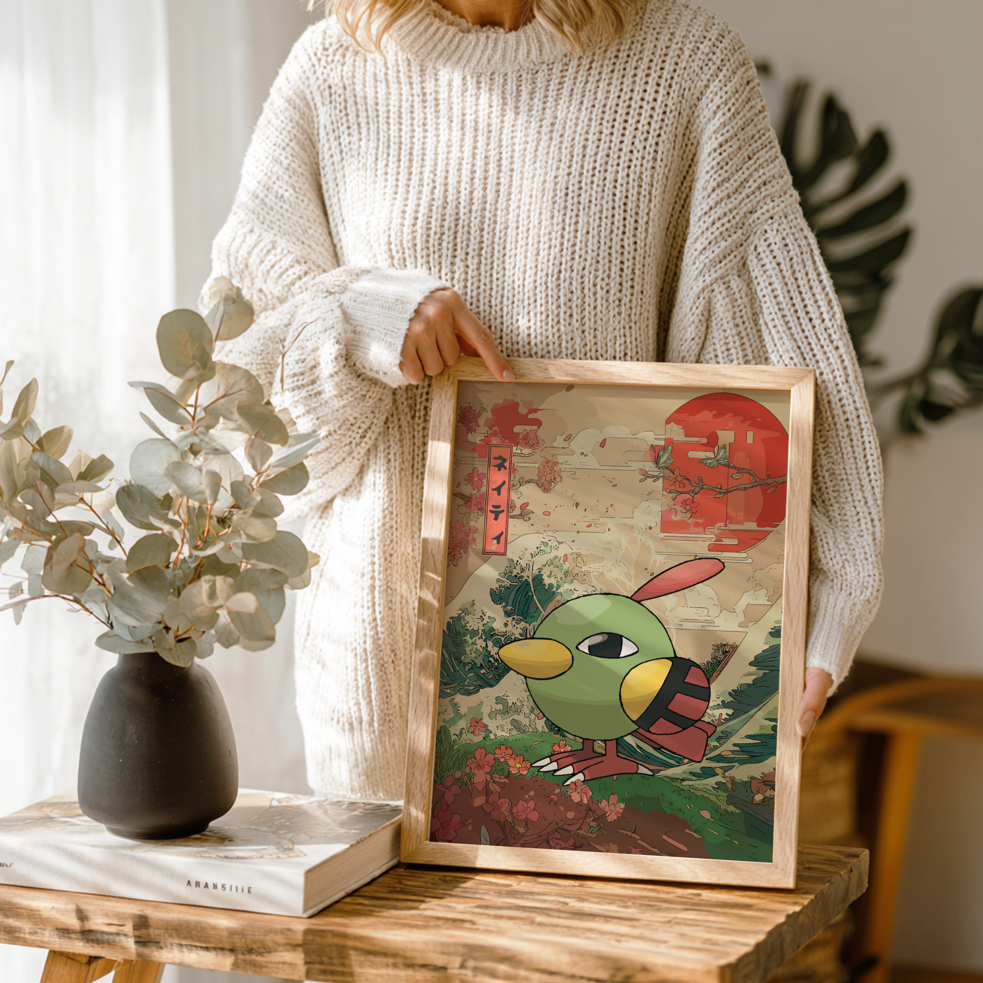 Person holding a framed artwork of Natu Pokemon with traditional elements on a wooden table.