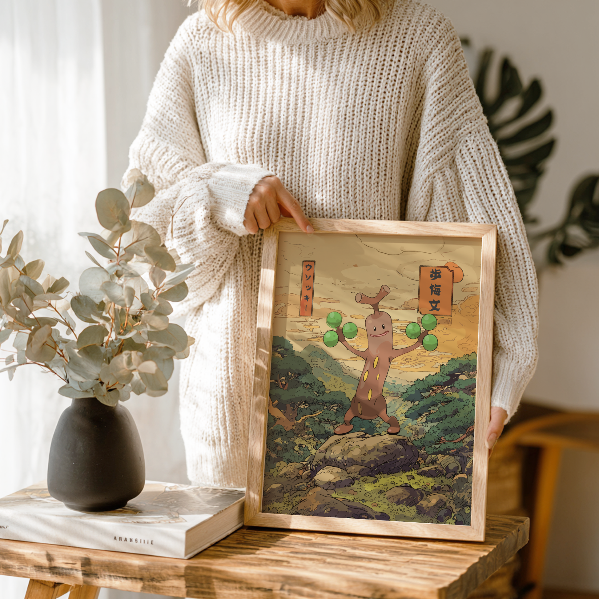 Person holding a framed artwork of Sudowoodo Pokemon in a natural setting, with a vase and book on a table.