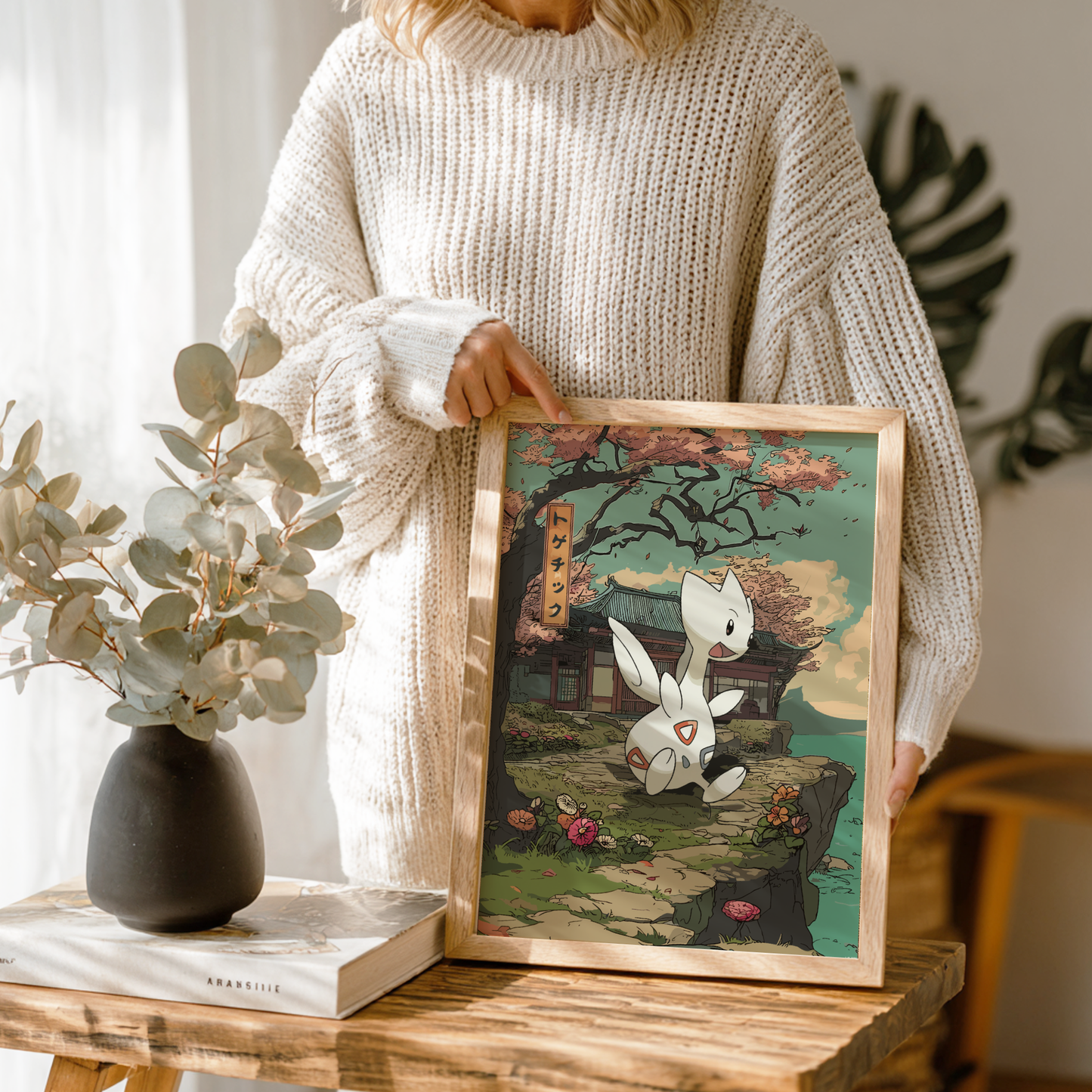 Person holding a framed artwork with Togetic Pokemon and cherry blossom design, standing next to a vase and book on a wooden table.