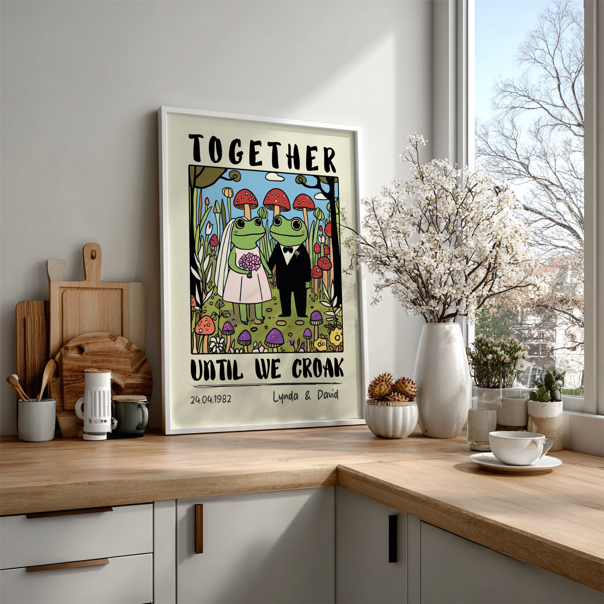 Framed artwork with frogs in wedding attire on a kitchen counter.