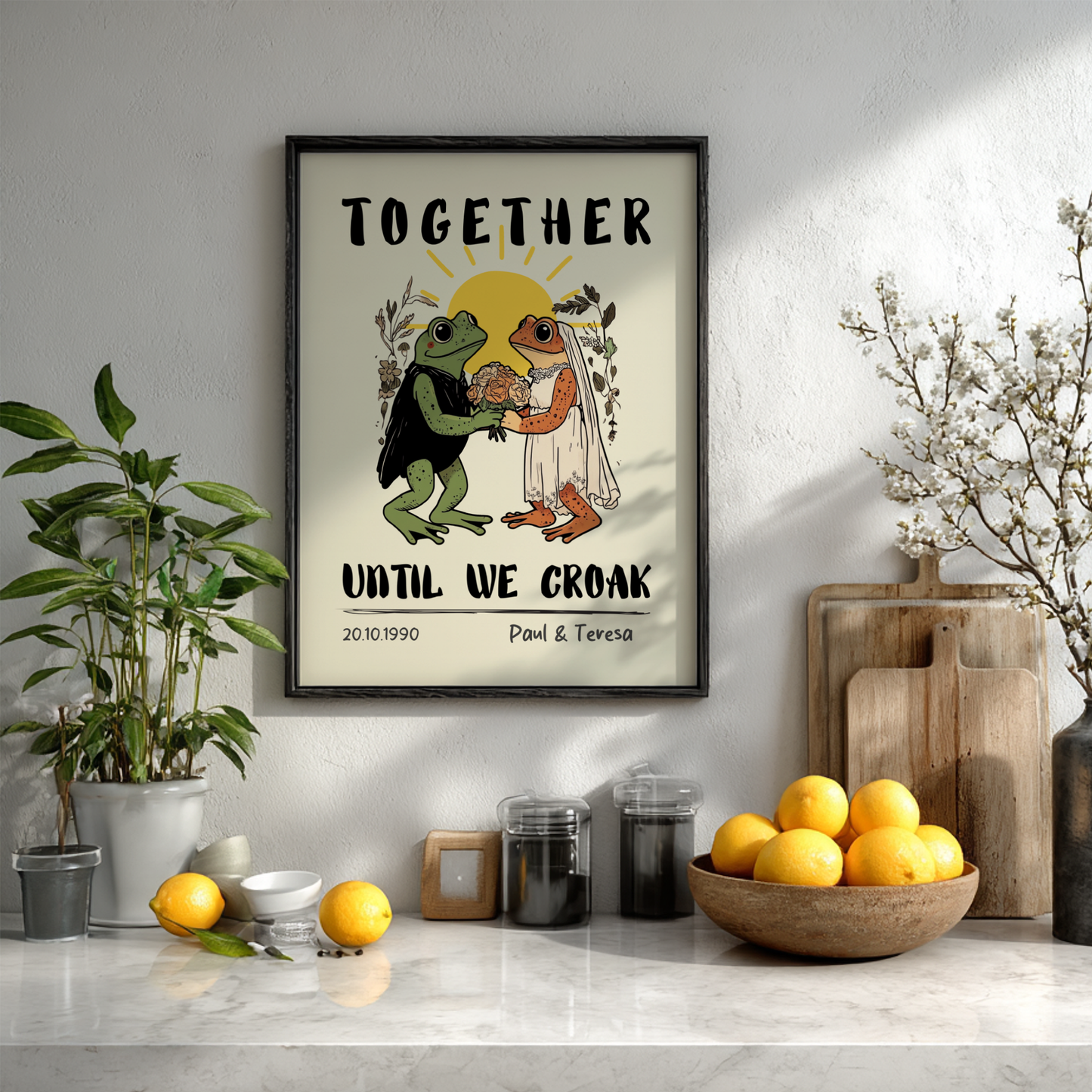 Framed artwork with frogs in wedding attire on a kitchen counter with fruits and plants.