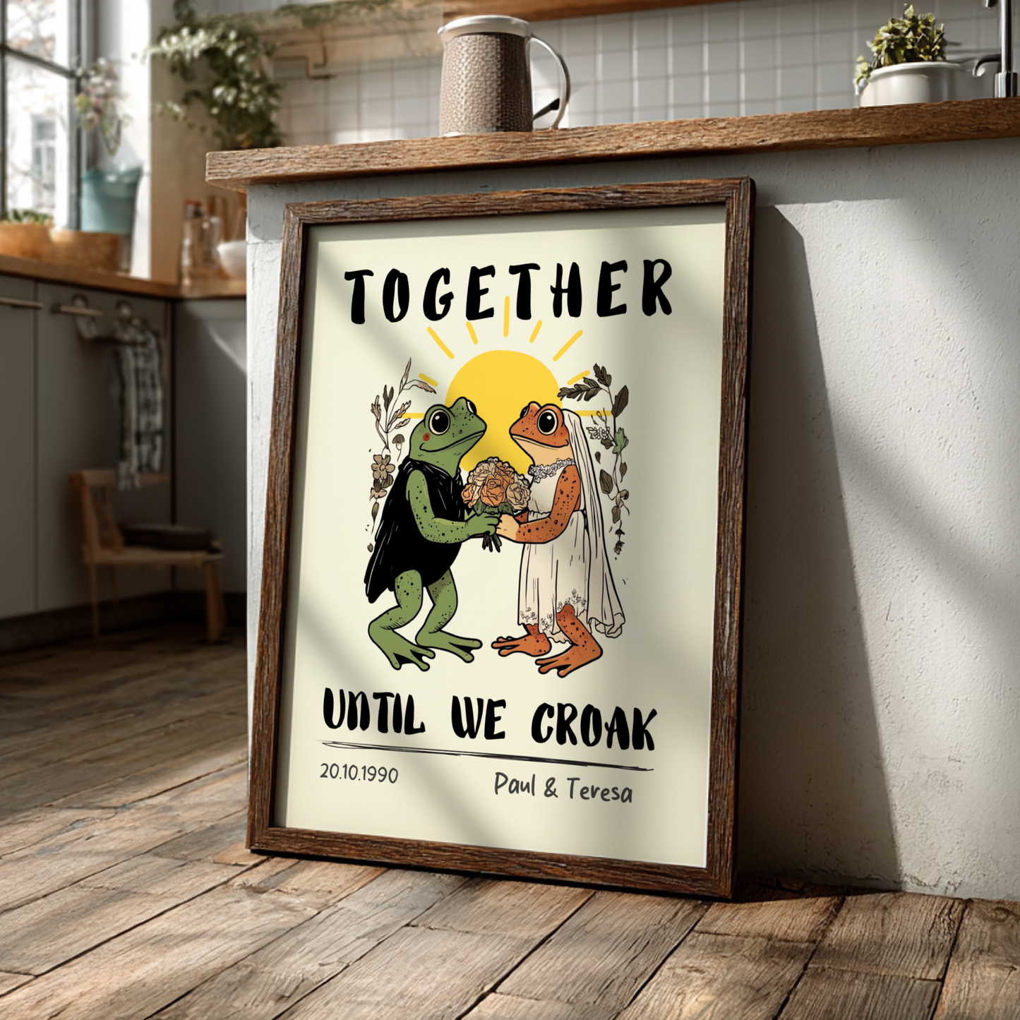 Framed artwork with two frogs holding hands and text 'Together until we croak' in a kitchen setting.
