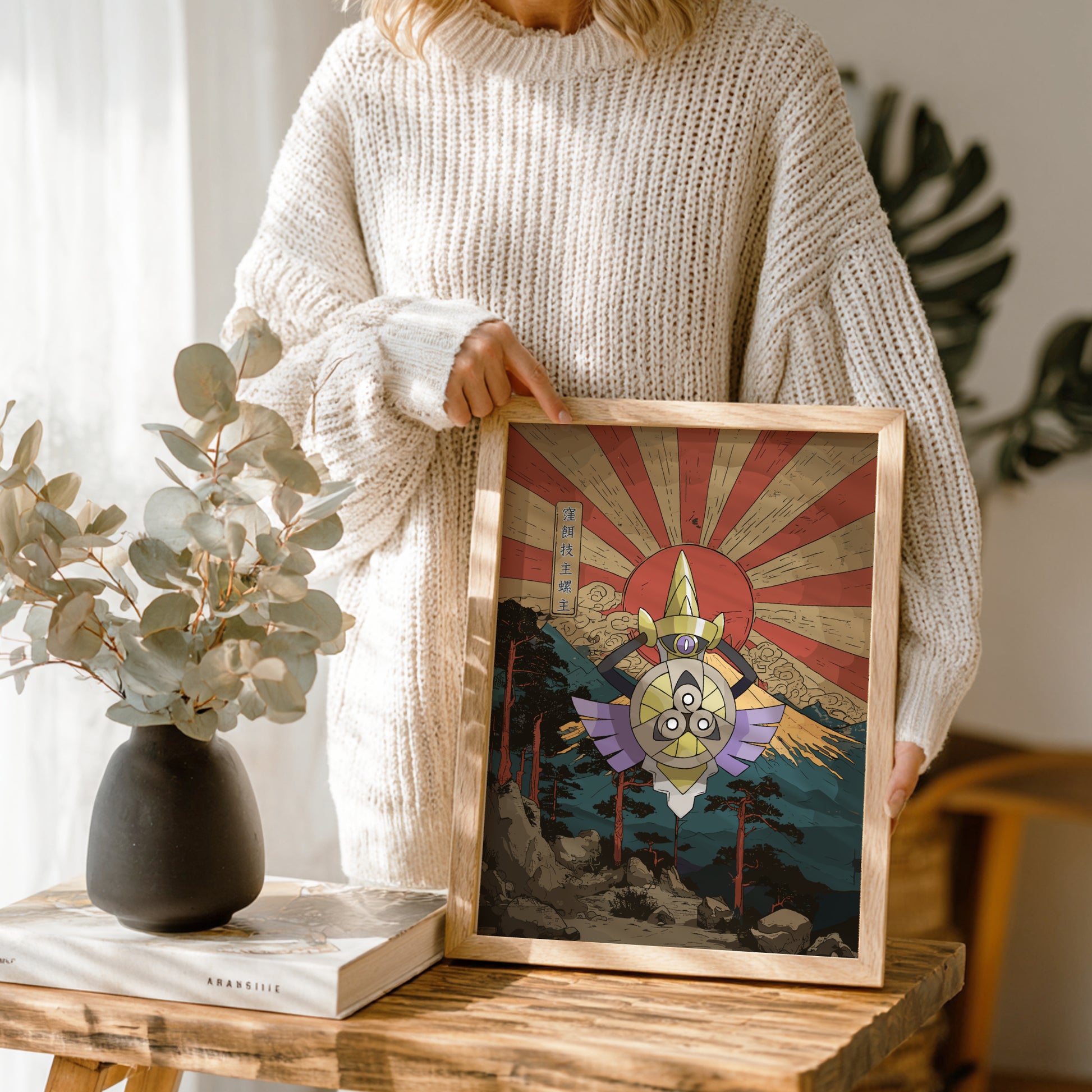 Person holding a framed artwork with a red sun and mountain landscape, standing next to a vase and books on a wooden table.