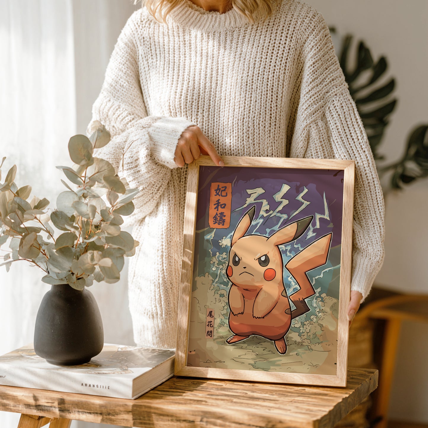 Person holding a framed artwork of an Angry Pikachu Pokemon on a wooden table with a vase and book in the background.