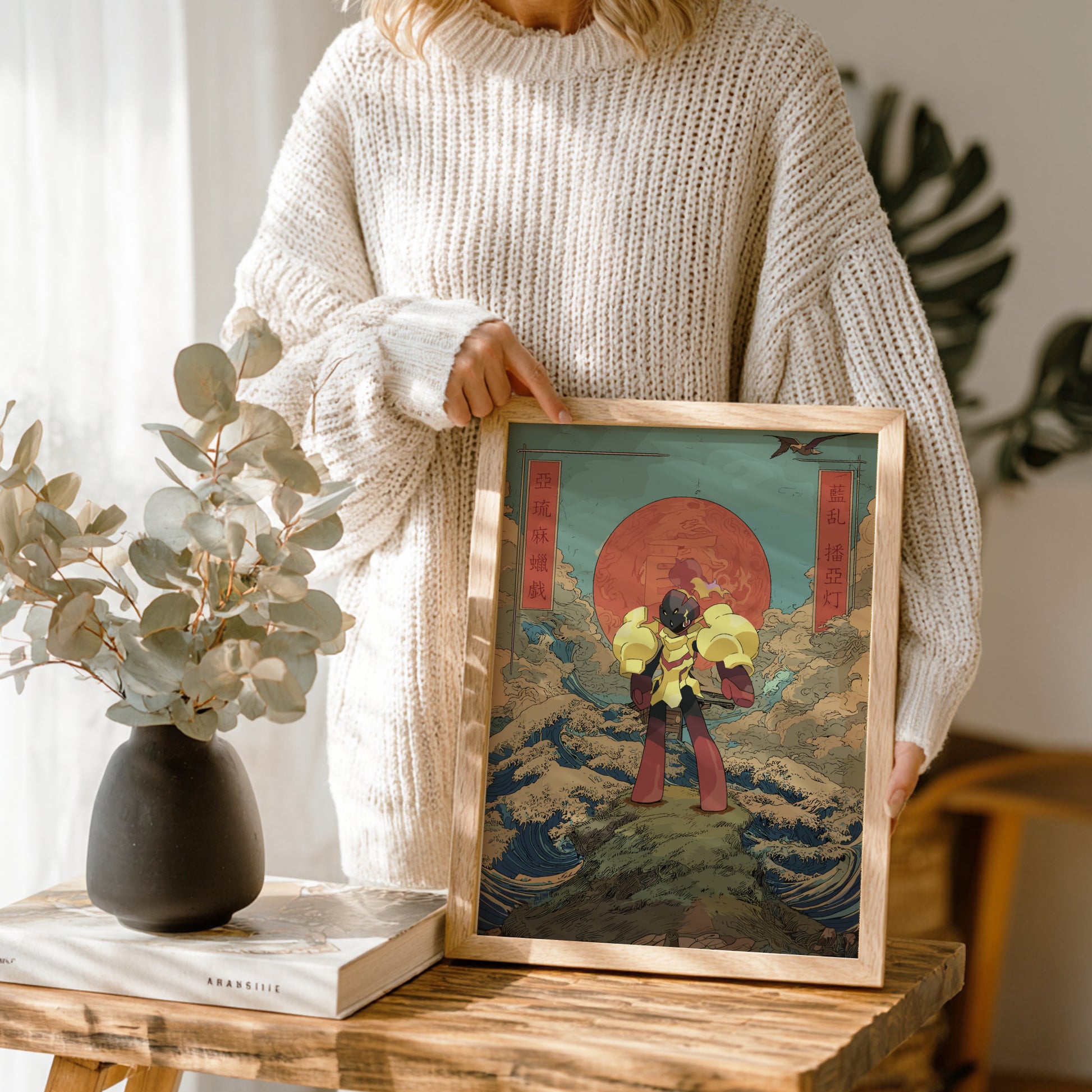 Person holding a framed artwork on a wooden table with a vase and books in the background.