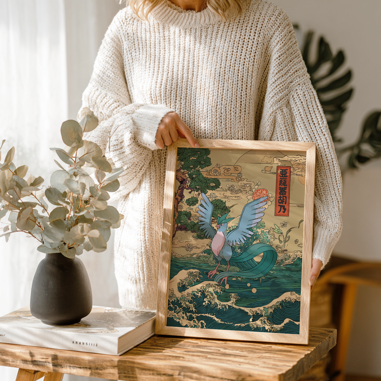 Person holding a framed artwork of Articuno Pokemon on a wooden table with a vase and book in the background.
