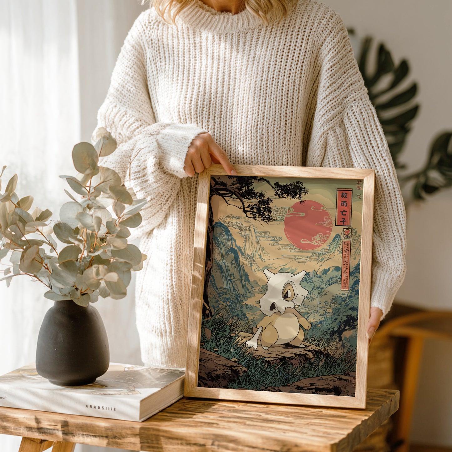 Person holding a framed artwork of Cubone Pokemon in a natural setting, with a vase and books on a table.