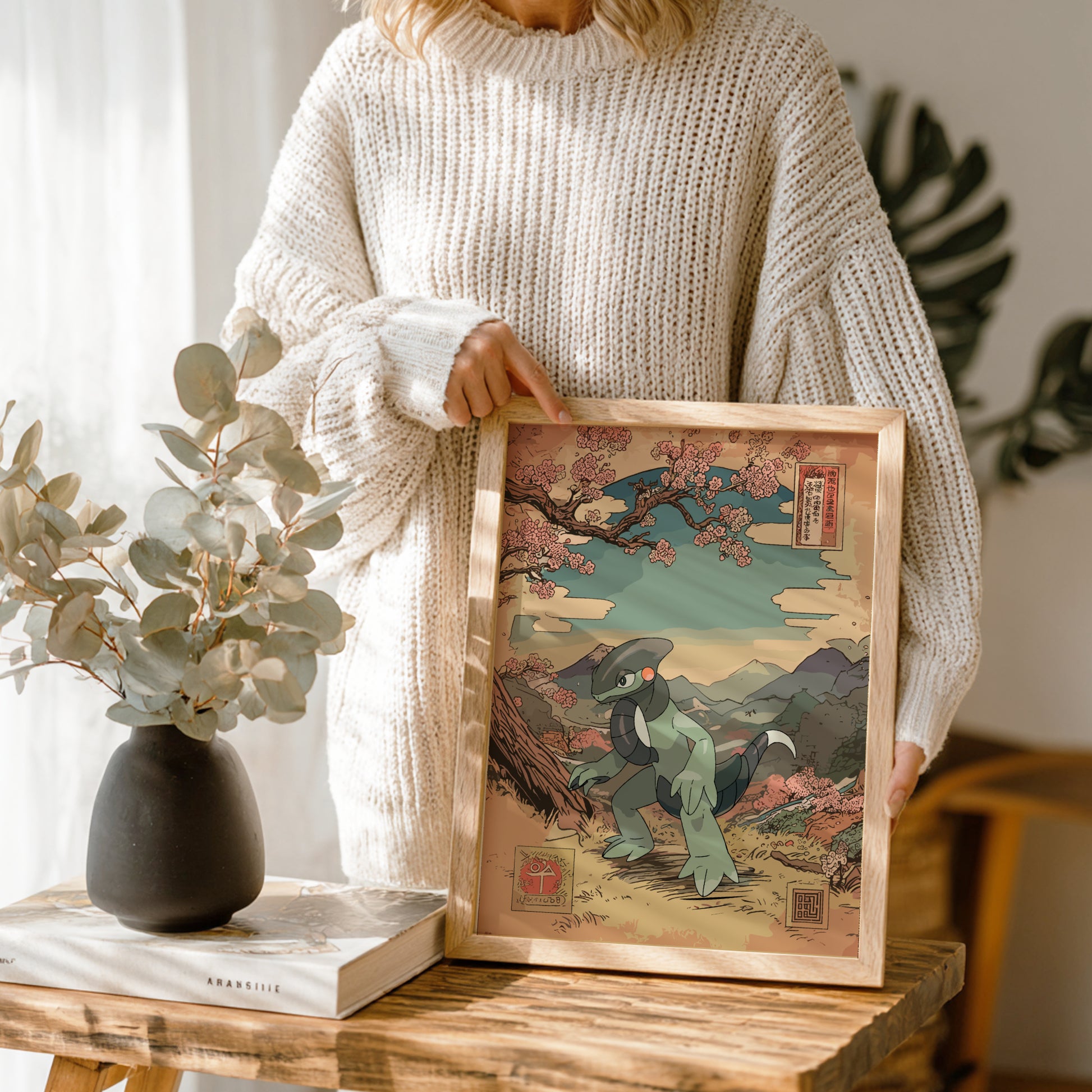 Person holding a framed artwork with a scenic design of Cyclizar Pokemon, standing next to a vase and book on a wooden table.