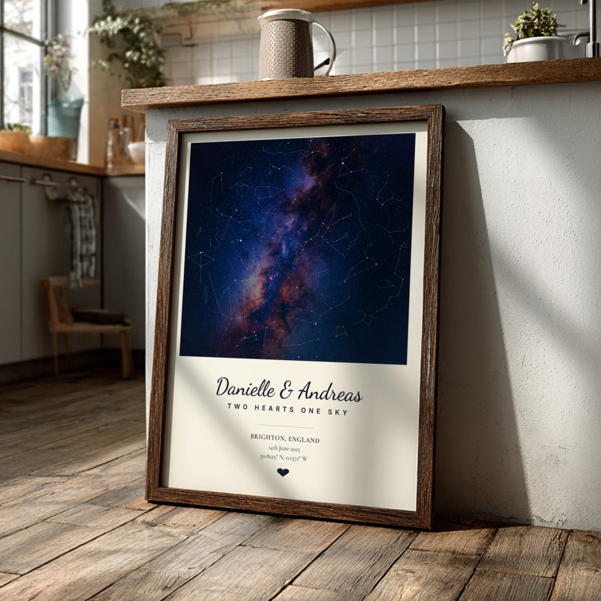 A framed personalized star map print leaning against a kitchen counter on a wooden floor. The print features a dark blue and purple galaxy night sky with constellation lines, a white lower section with the names 'Danielle & Andreas' and 'Two Hearts One Sky' in script, and a rustic brown wood frame.