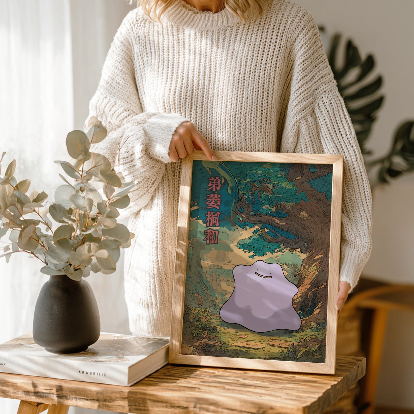 Person holding a framed artwork with Ditto Pokemon in a forest scene, standing next to a vase and books on a wooden table.