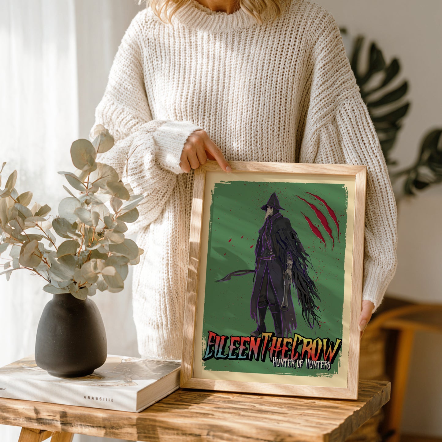 Person holding a framed artwork of Eileen The Crow from Bloodborne on a wooden table.