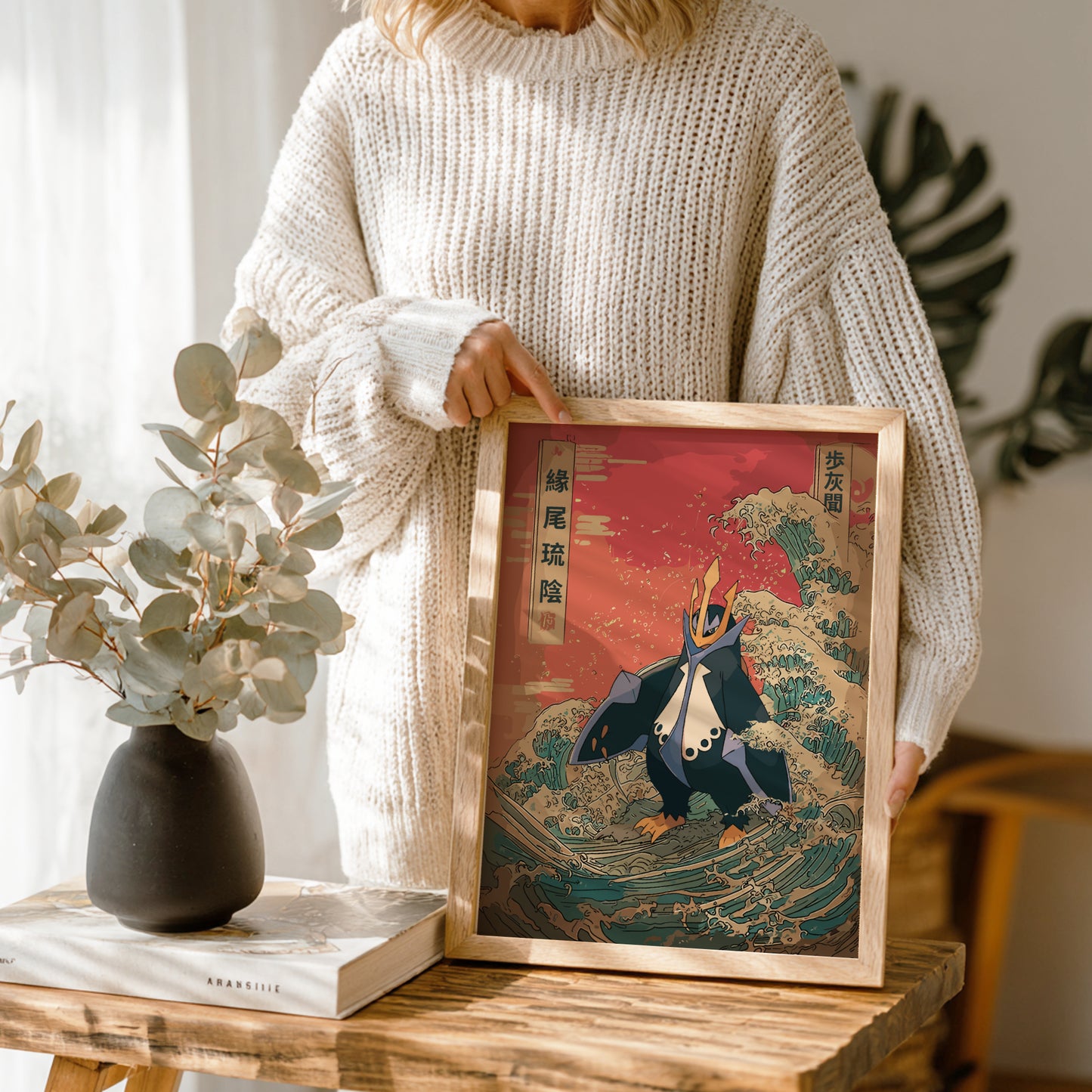 Person holding a framed artwork of Empoleon Pokemon character on a wave, with a vase and book on a table in the foreground.