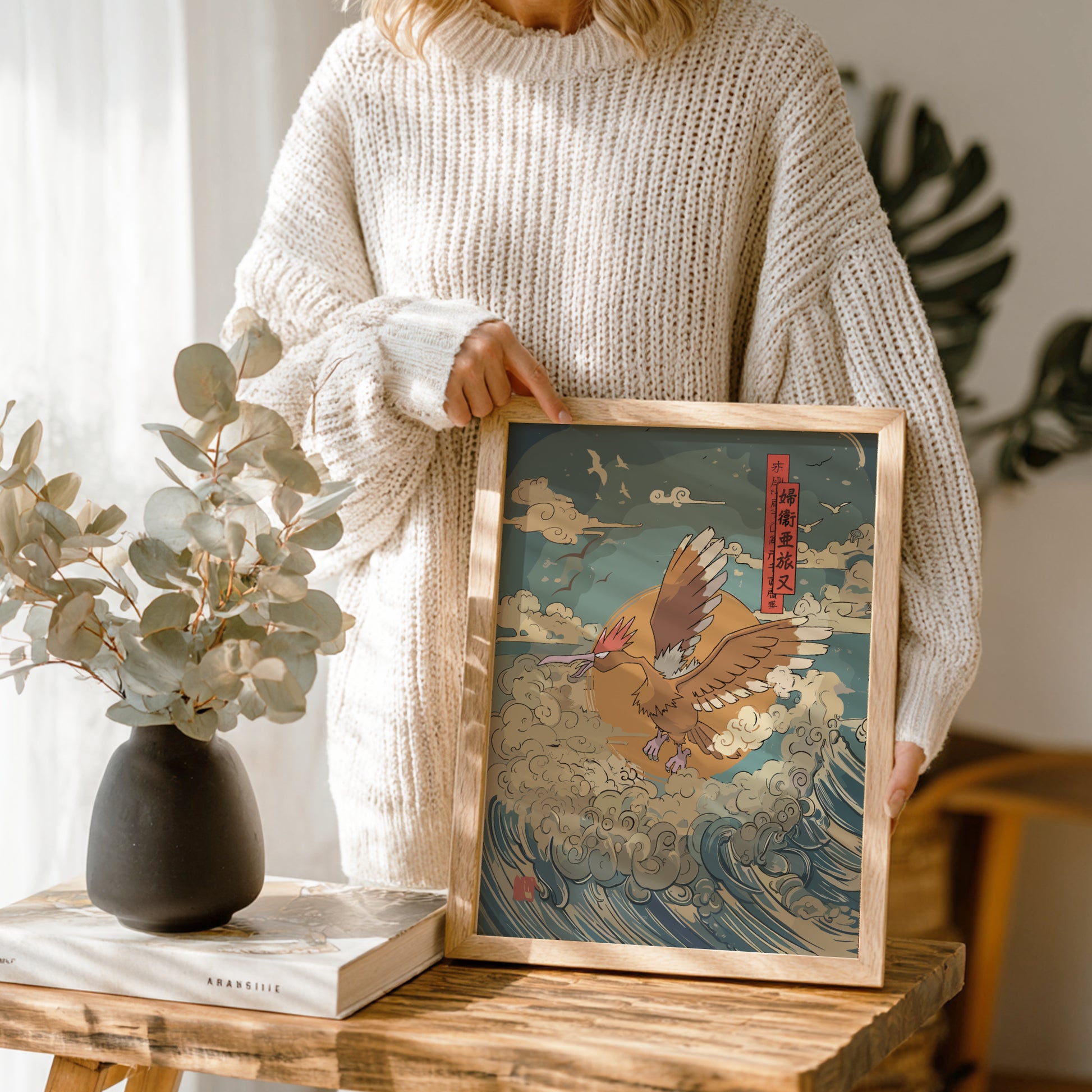 Person holding a framed artwork of Fearow Pokemon on a wave, with a vase and book on a wooden table.