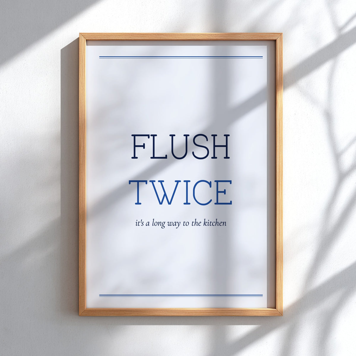 A framed typographic wall art print reading “FLUSH TWICE” with smaller text “it’s a long way to the kitchen,” shown in a light wood frame against a white wall with soft shadow overlays.