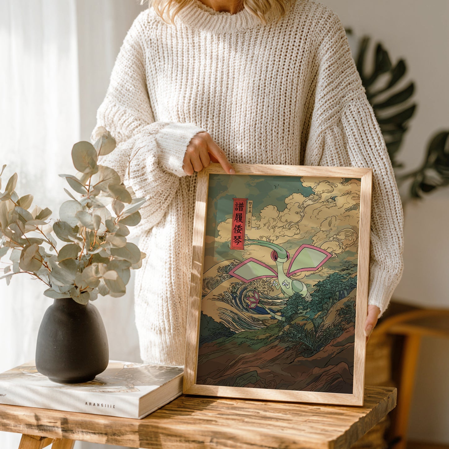 Person holding a framed artwork on a wooden table with a vase and book in the background.