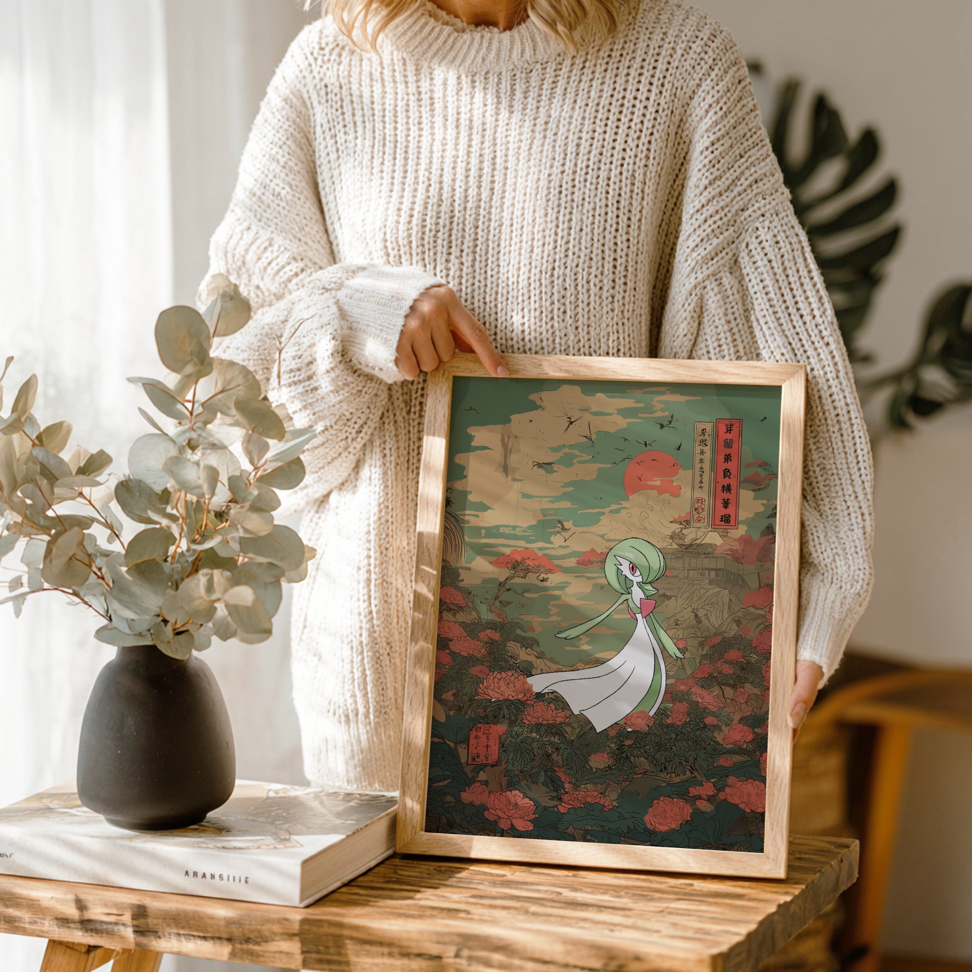 Person holding a framed artwork on a wooden table with a vase and book in the background