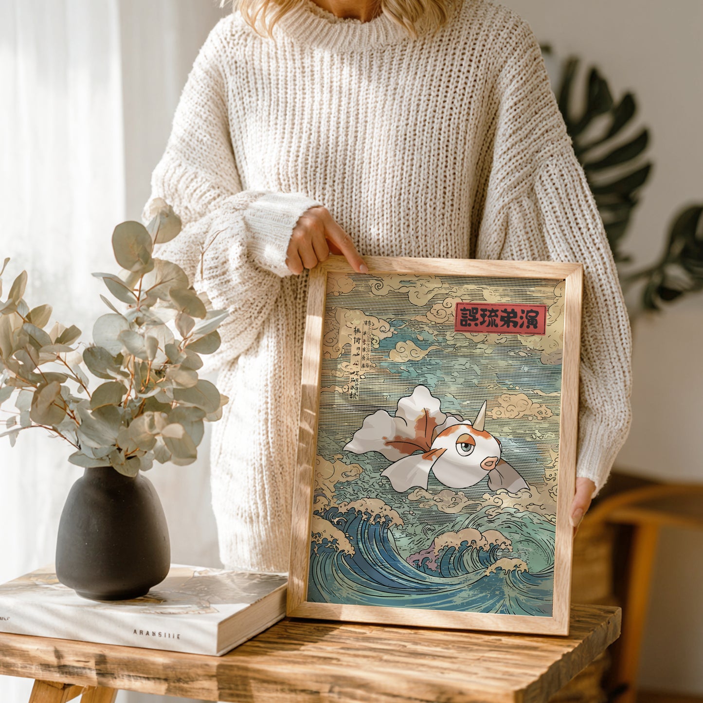 Person holding a framed artwork of Goldeen Pokemon in waves on a wooden table with a vase and book in the background.