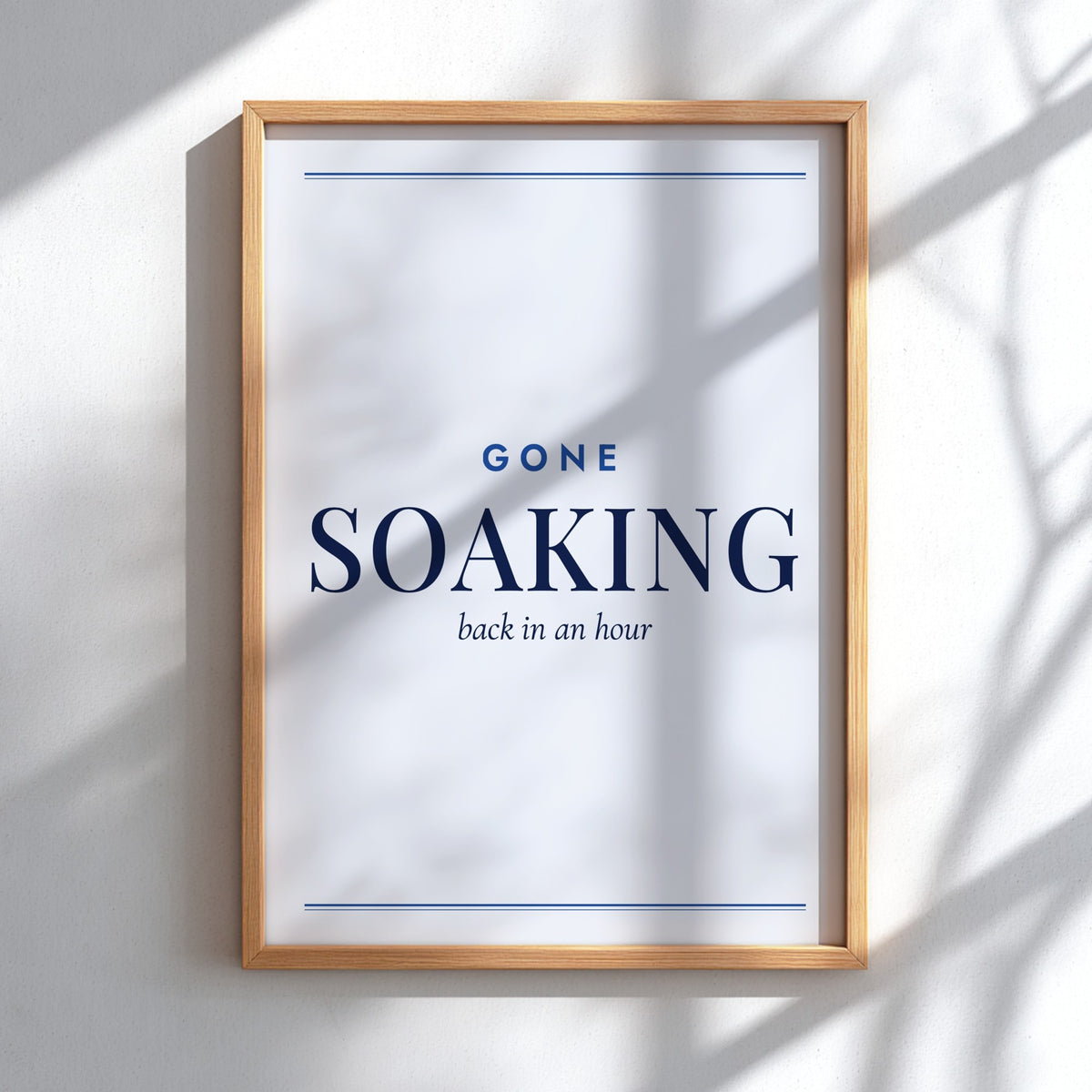 A framed wall art print with the words 'GONE SOAKING back in an hour' in navy text on a white background, mounted on a light wall with soft shadow overlays.
