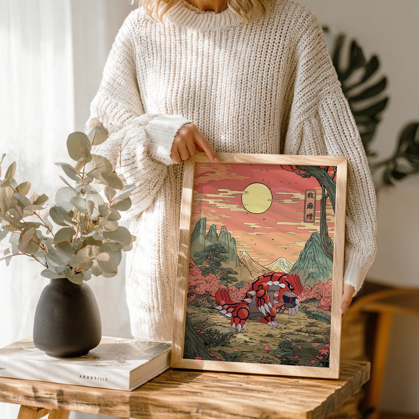 Person holding a framed artwork with a mountain and Groudon Pokemon design, standing next to a vase and books on a wooden table.