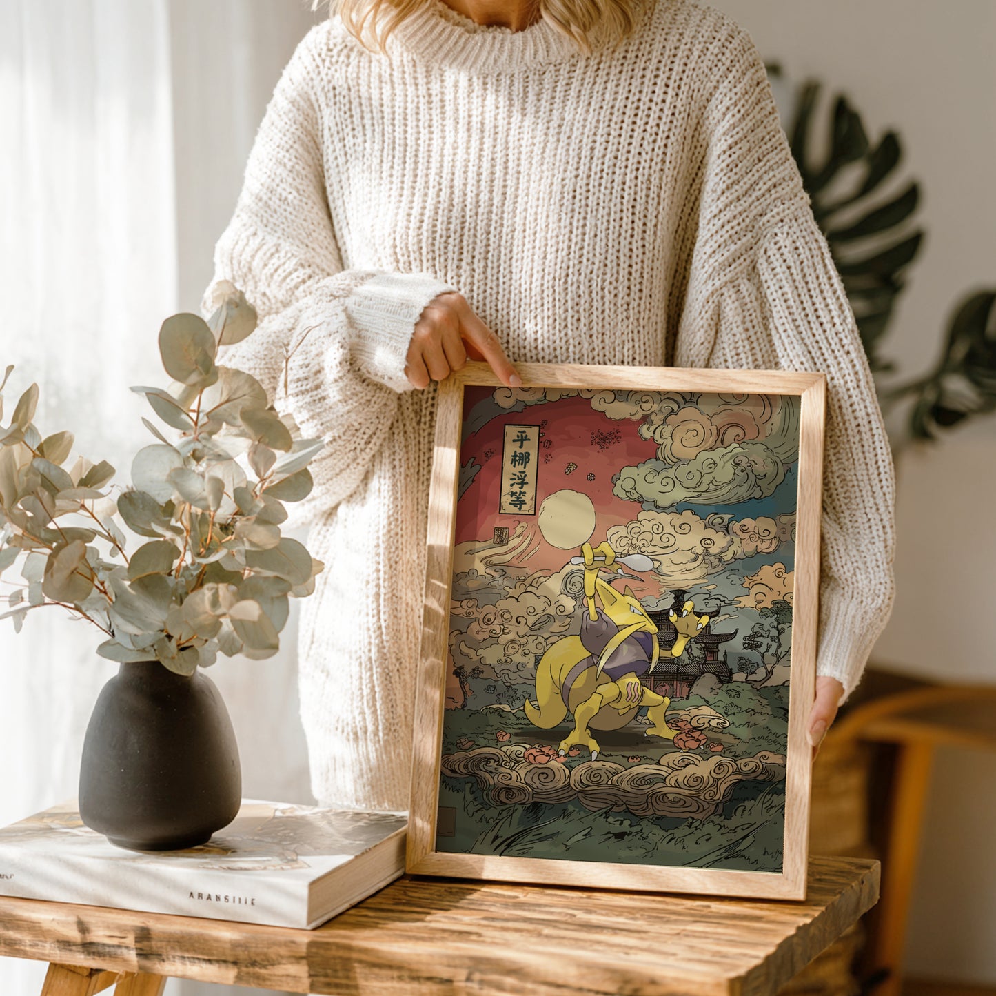 Person holding a framed artwork on a wooden table with a vase and book in the background.