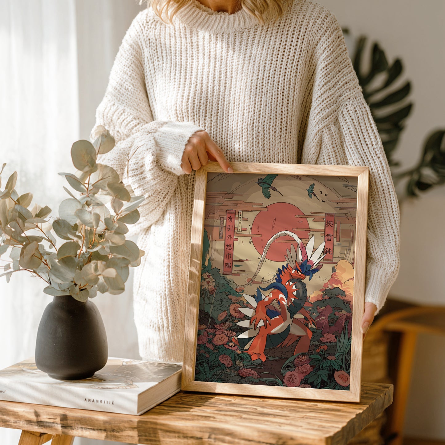 Person holding a framed artwork with a nature-themed design of Koraidon Pokemon, standing in a room with plants and decor.